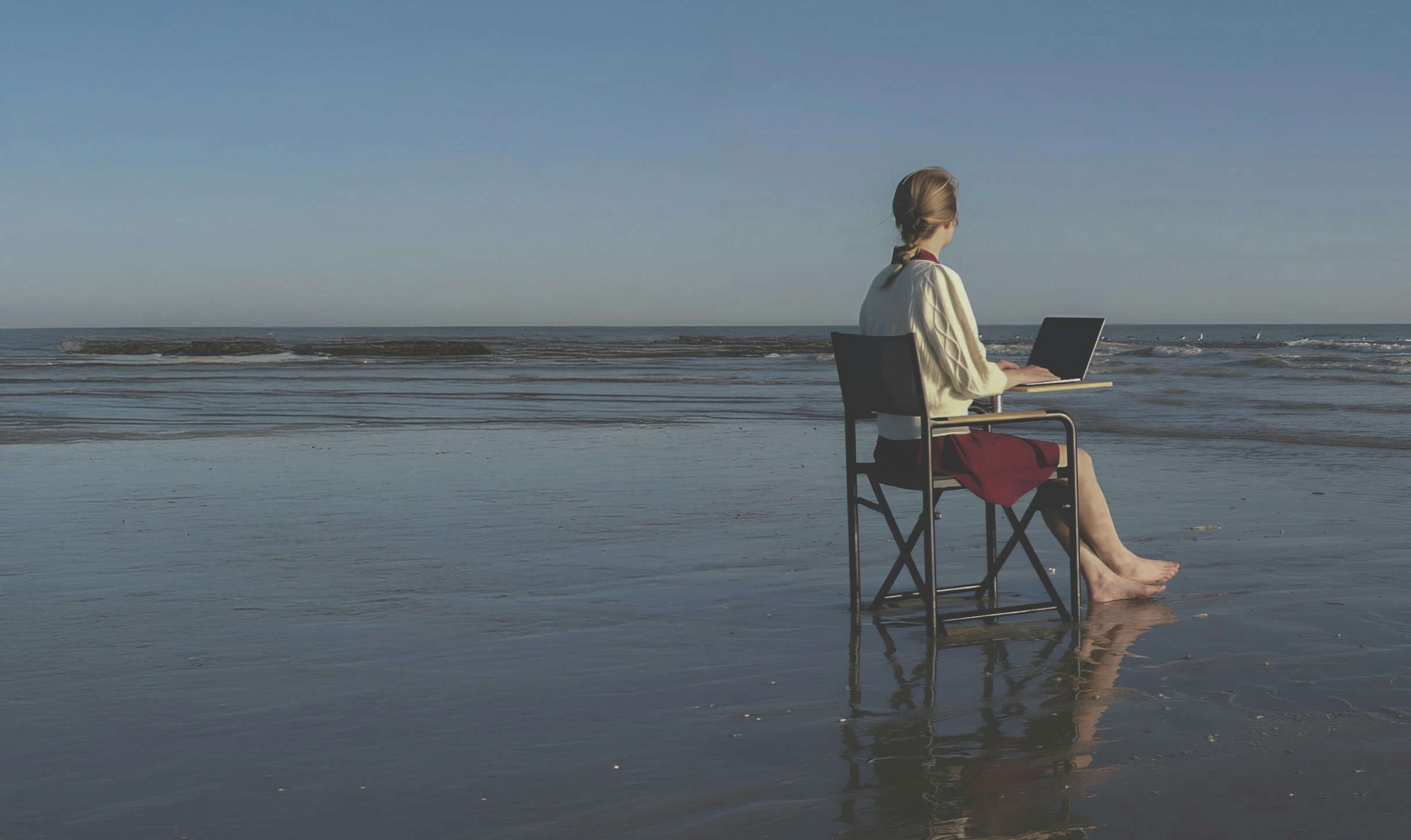 The image shows a woman sitting on a folding chair at the edge of a beach, using a laptop on a small desk. Her feet are bare, touching the wet sand, and the ocean stretches out in front of her under a clear blue sky. The scene captures a peaceful, remote work environment, symbolising flexibility, work-life balance, and the ability to work from unconventional locations. This visual is ideal for websites discussing remote work, digital nomad lifestyles, or hybrid work models.