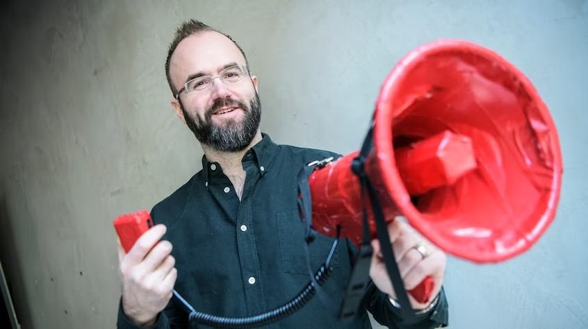 Torso shot of Magnus Larrson, a white man wearing glasses and a dark button-up shirt, holding a red megaphone.