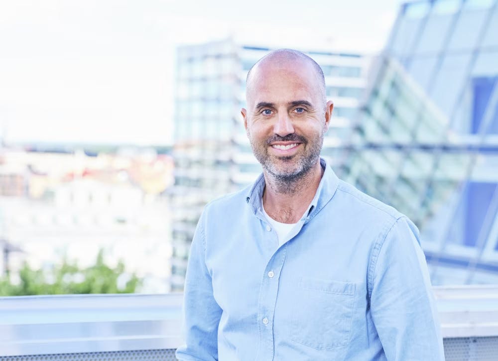 Upper-body shot of Erik Olofsson, a white man with a beard. He wears a light blue button-up shirt and stands outside during the day, in front of a glass building.