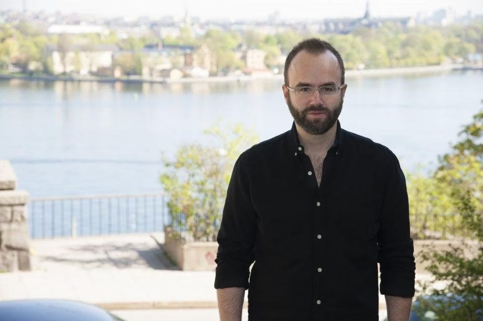 Magnus Larsson, a white man wearing a black shirt and glasses, standing in front of a lake during the day.