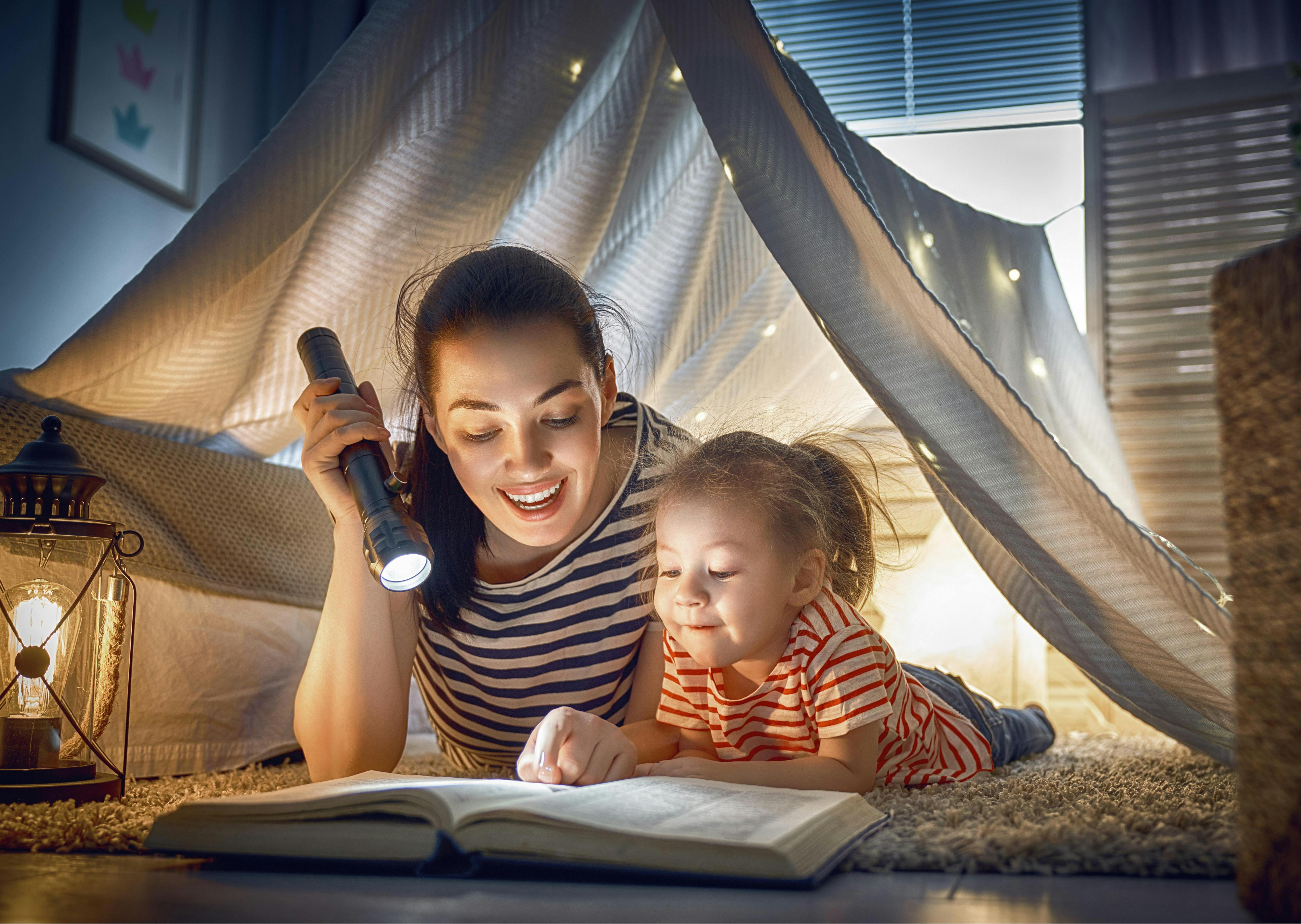 A mother reading a book with her child under a tent made out of a blanket