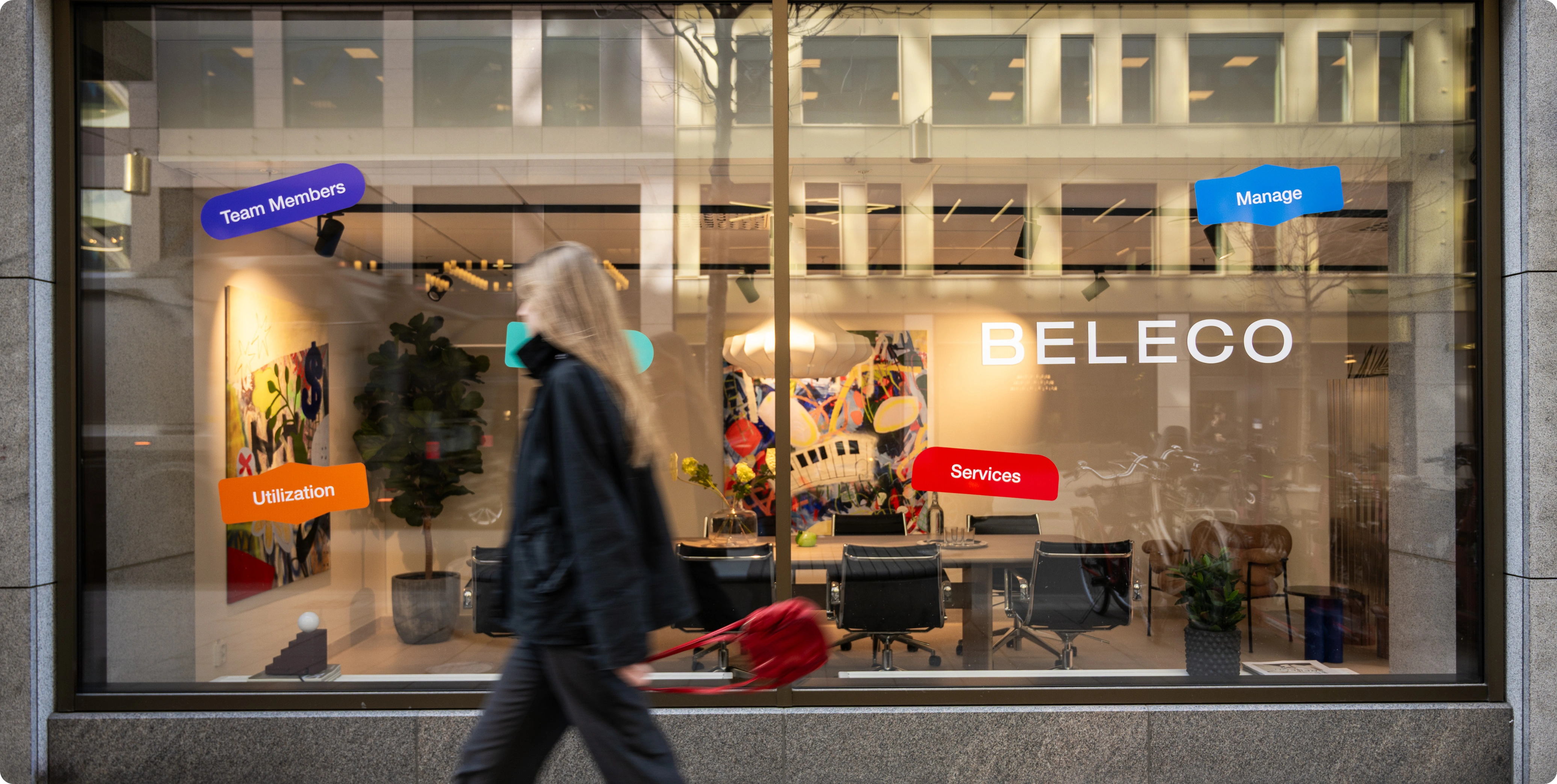 Facade view of Beleco’s showroom on Malmskillnadsgatan with large glass windows. Inside, a meeting table, artworks, and design furniture are visible. A person walks past on the sidewalk.