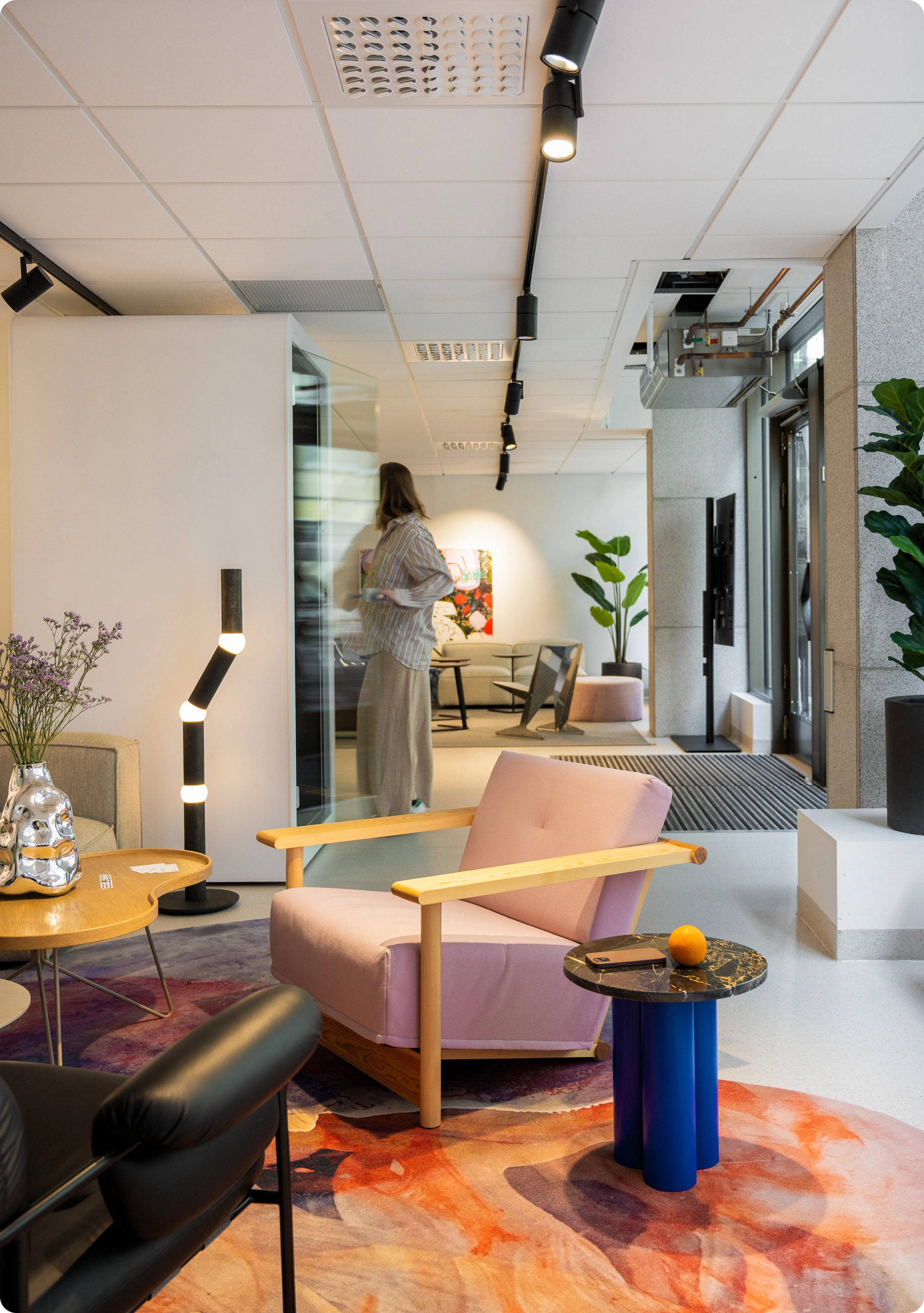 Lounge area with sofas, chairs, rugs, and plants. A person walks by.