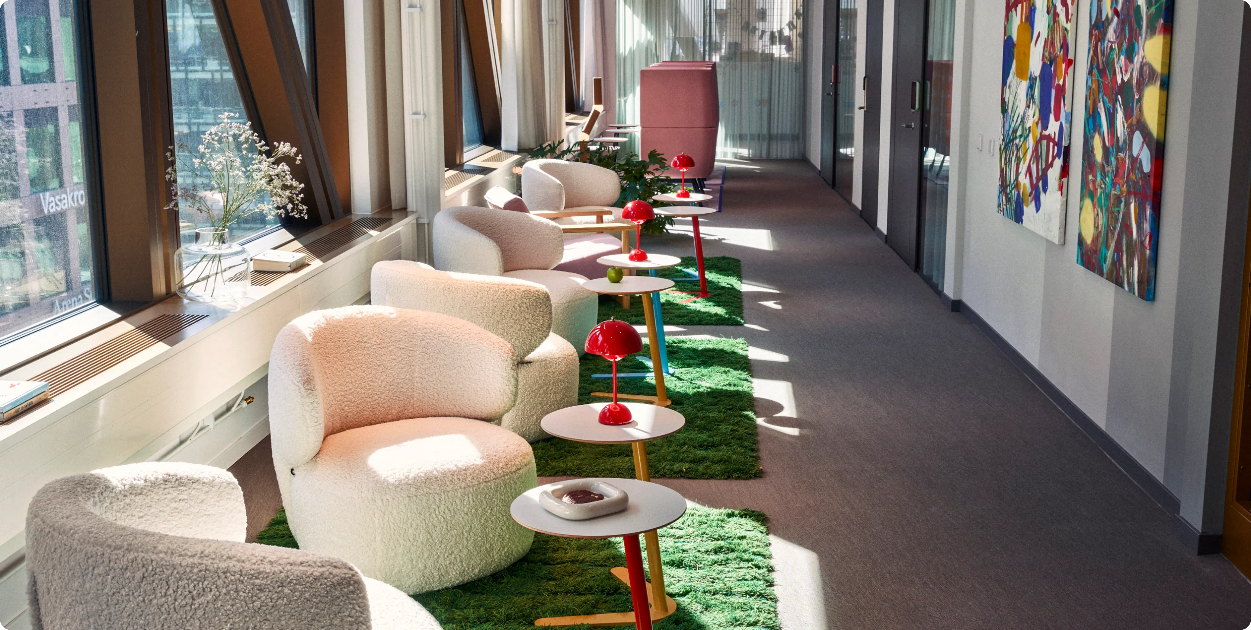 Lounge area at King’s Stockholm office with white armchairs, small tables with red lamps, green rugs, and colorful artwork on the walls.