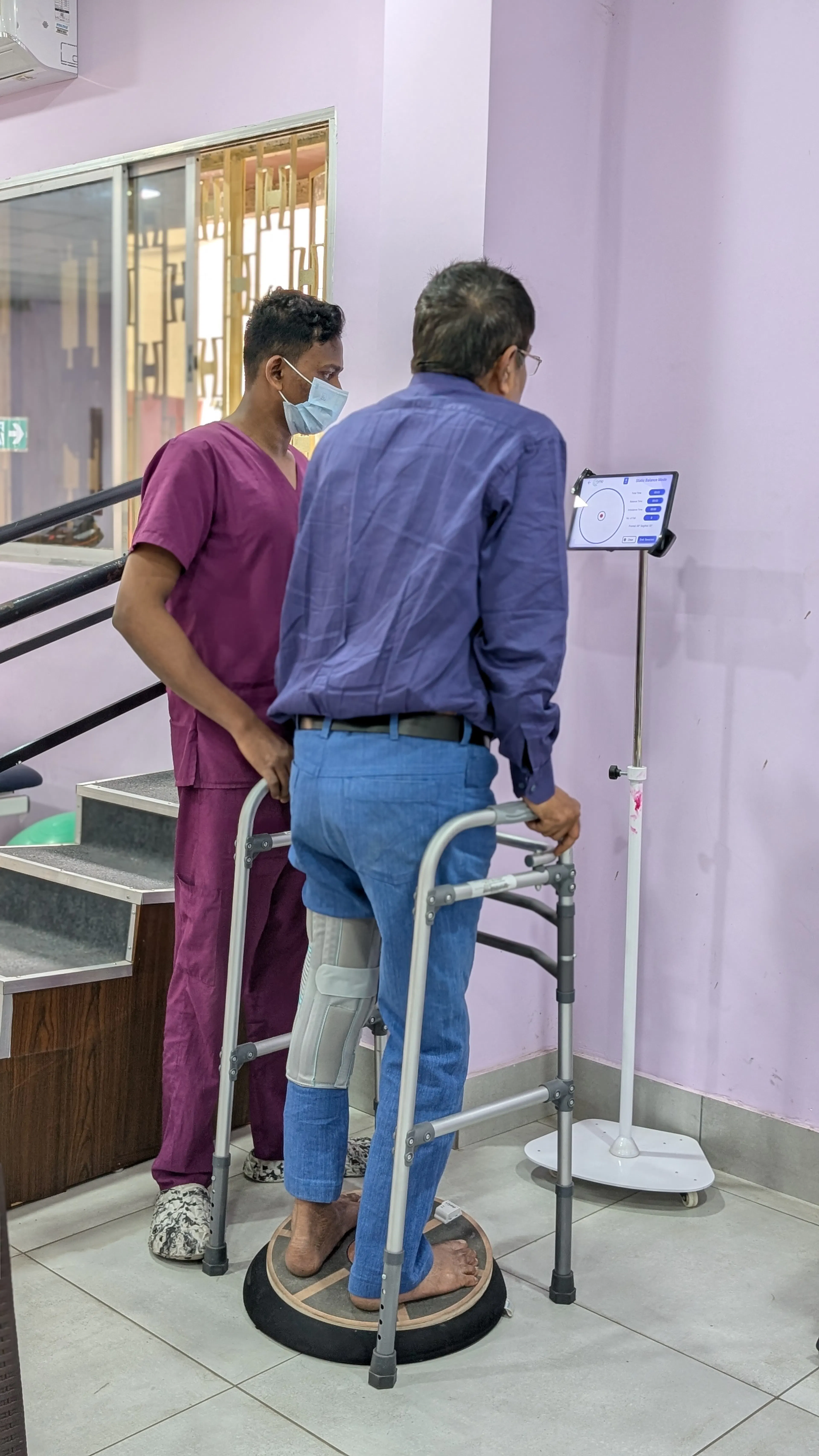 Patient with a walker standing on a balance platform with a screen in front, practising balance with a therapist close by in VR-style rehab