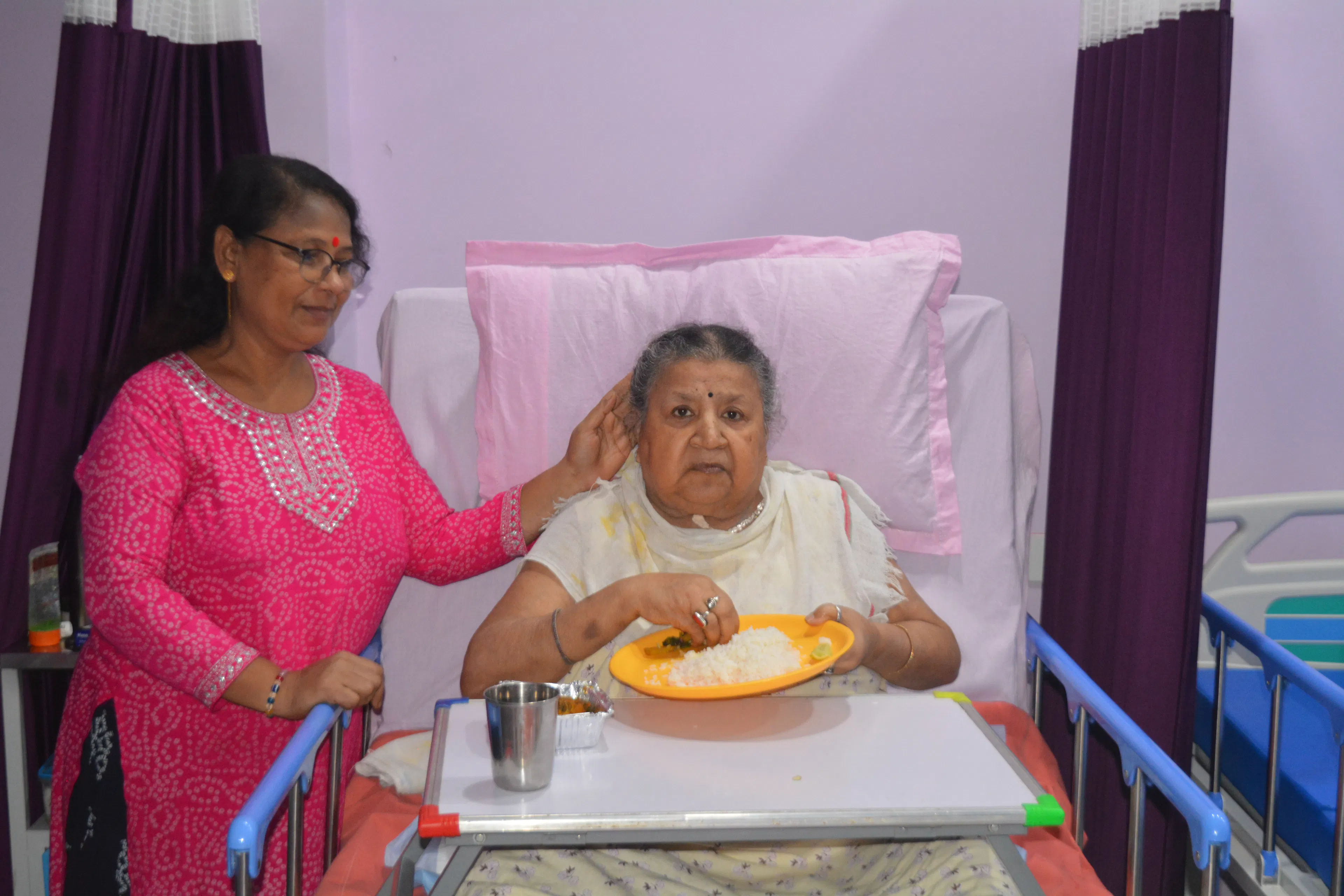 Elderly woman feeding herself from a plate on an over-bed table while a caregiver stands beside her