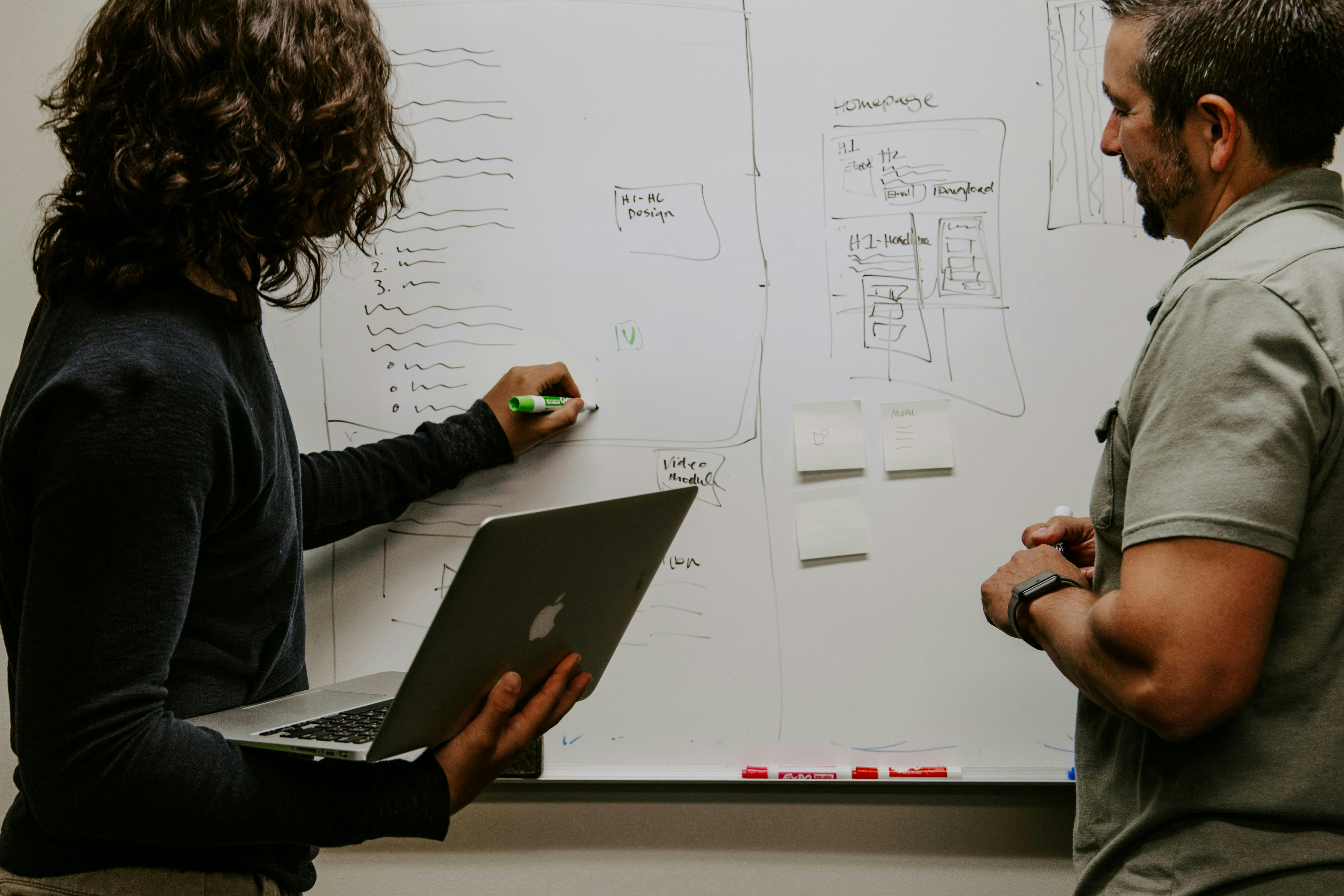 Two people are collaborating in front of a whiteboard. One person is holding a laptop and writing on the board with a green marker, while the other stands nearby, listening and observing. The whiteboard is filled with sketches, diagrams, and notes.