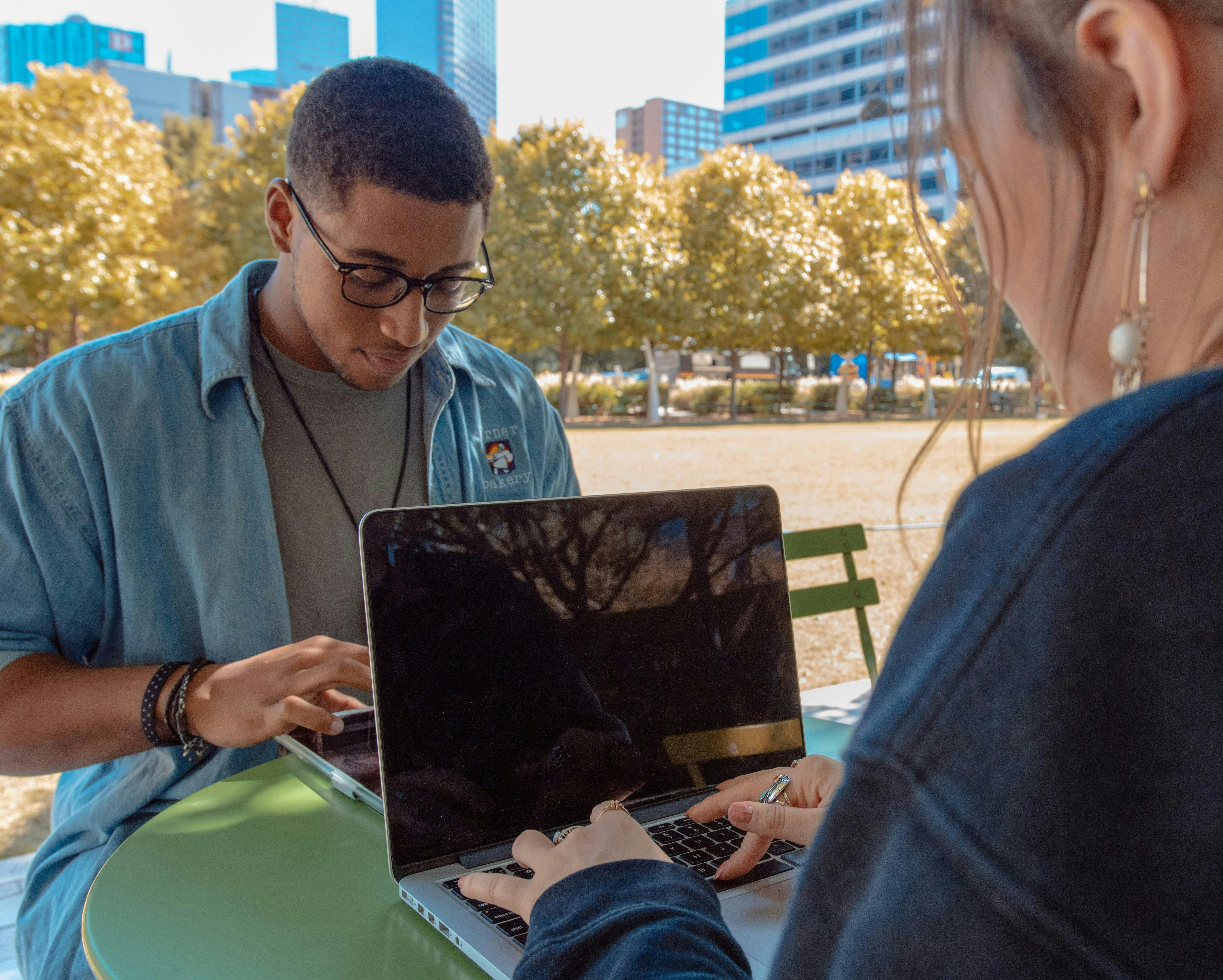 Two young  people working on laptops at an outdoor table in a sunny park, with trees and buildings in the background.