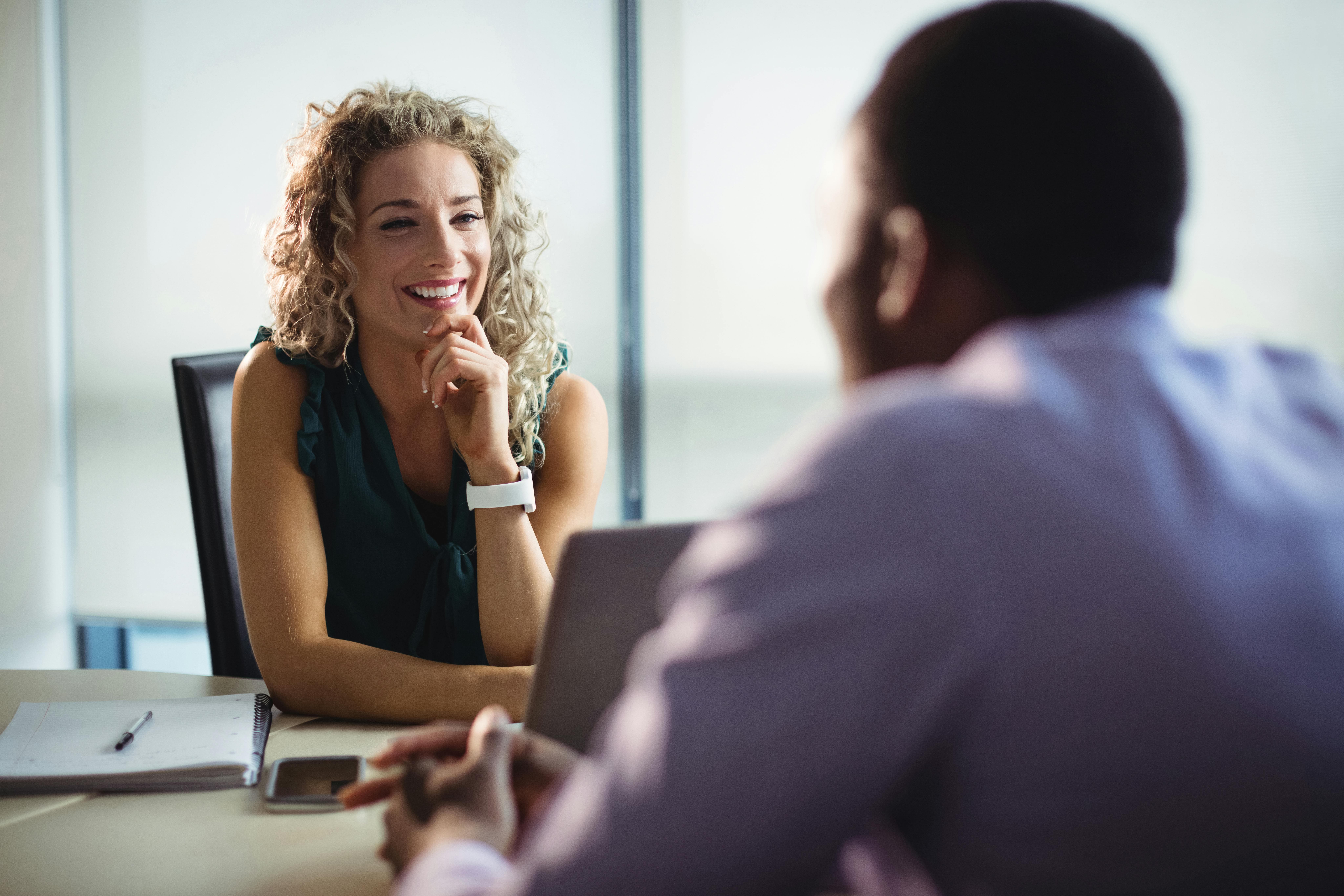 A smiling woman with curly hair sits at a desk in a bright office, leaning forward and listening attentively while talking with another person across from her who is using a laptop.