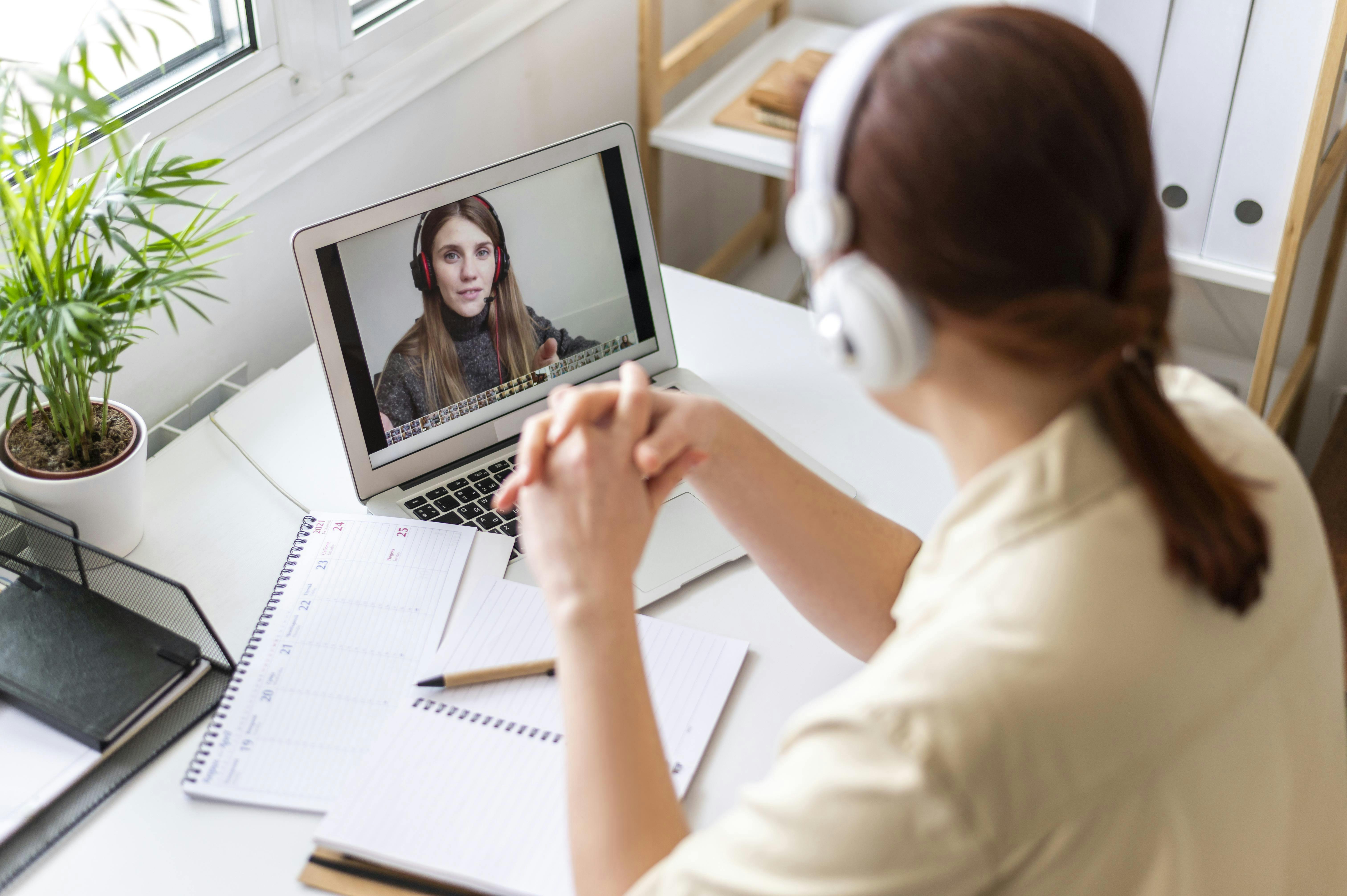 Person sitting at a desk wearing headphones, taking part in a video call on a laptop while writing notes in a notebook, in a bright home office with a plant nearby.