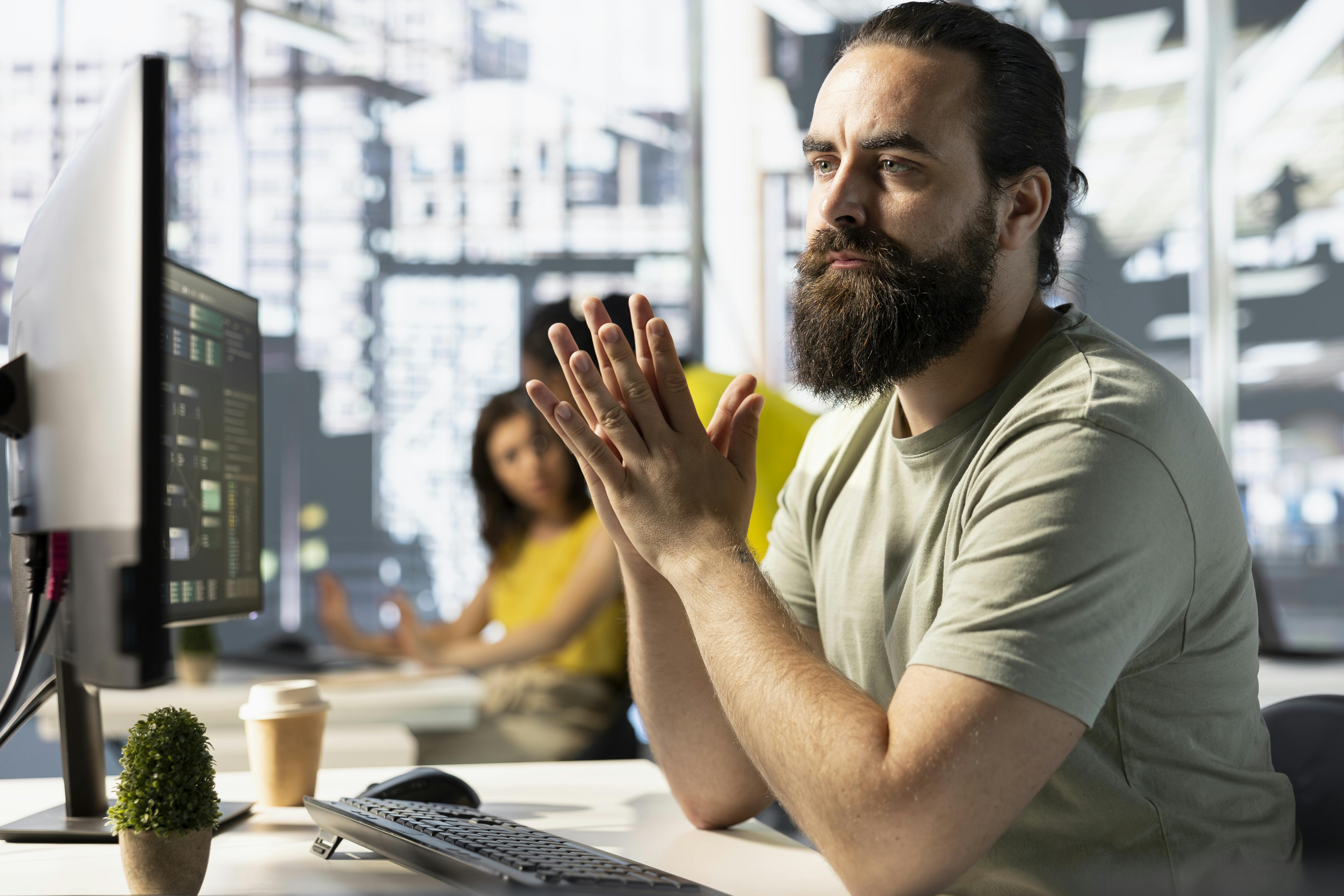 A bearded man in a casual green shirt is seated at a desk, focused on a computer screen, hands clasped together. A blurred figure in yellow is seen in the background.