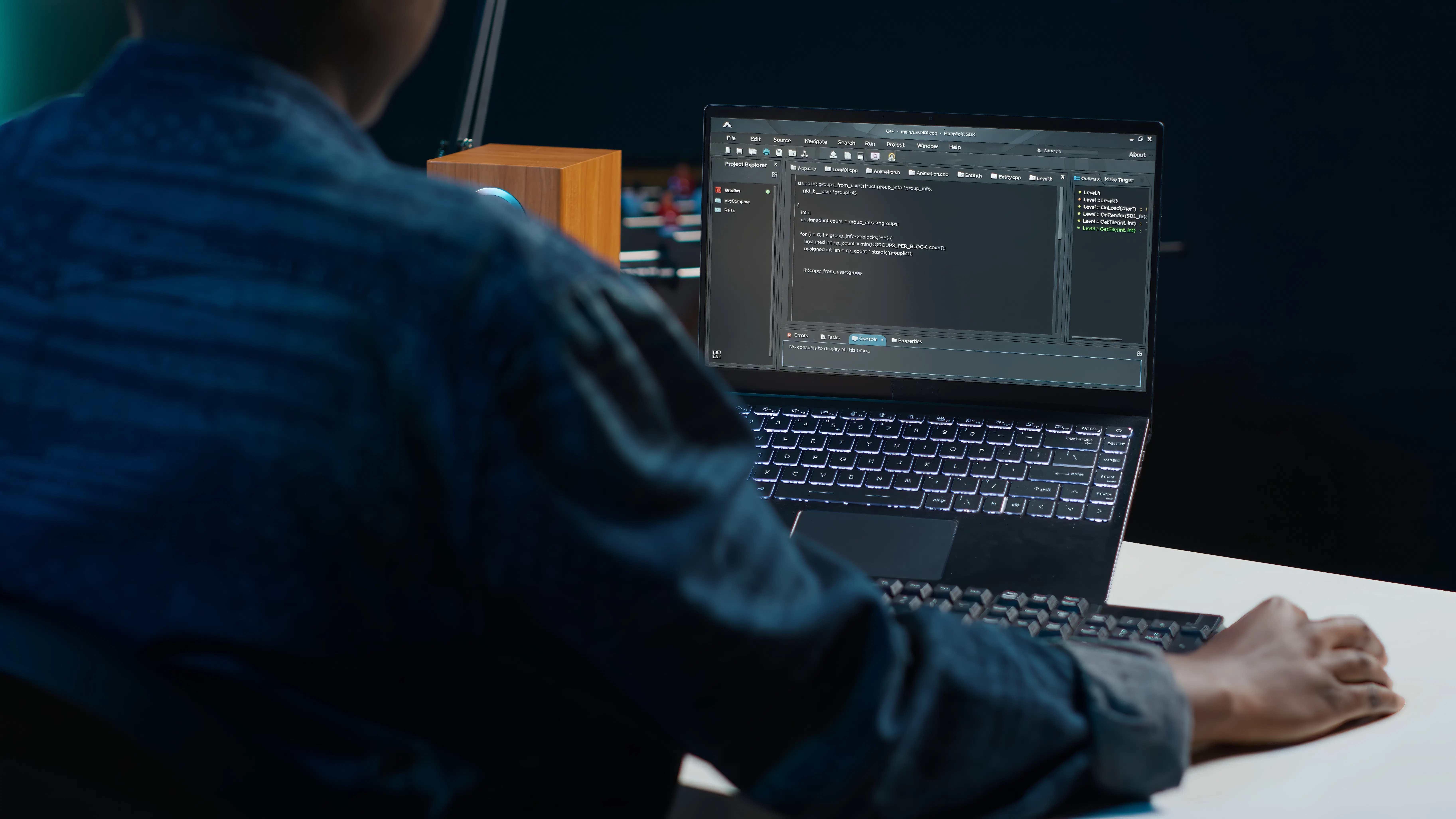 A person sits at a desk using a laptop, displaying code on a dark-themed interface. The setting is dimly lit, suggesting focus and concentration.