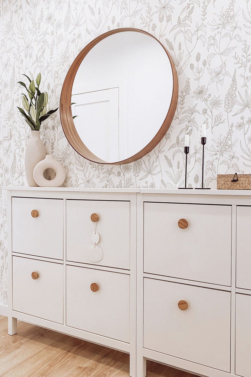 weiße schmale Kommode im Flur, runder Spiegel in Holzrahmen, einfarbige Tapete mit floralen Mustern