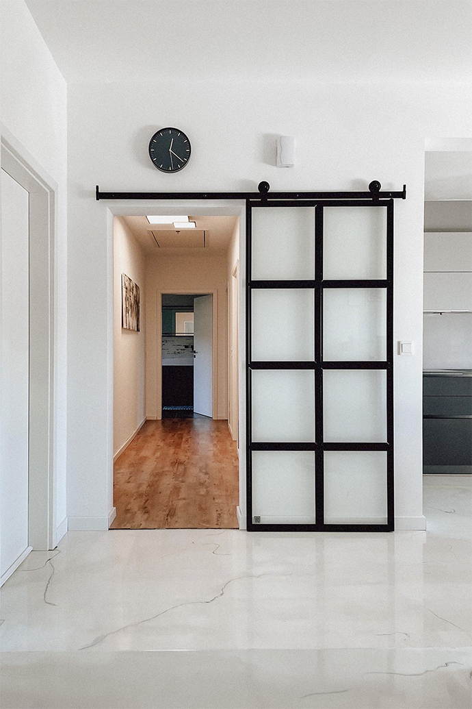 Glasschiebetür, Glastür trennt den Flur, heller Marmorboden, weiße Wände und schwarze Accessoires, schwarze Uhr an weißer Wand, Metalltür mit Glas