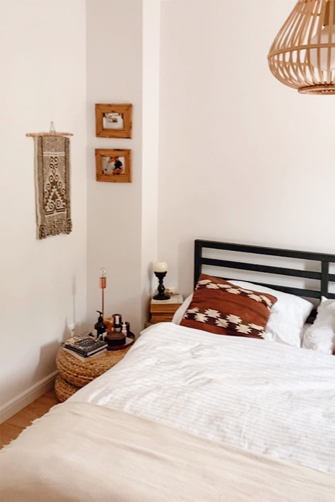 Weiße Wände in einem natürlichen Schlafzimmer, Boho-Dekorationen an einer weißen Wand im Schlafzimmer, ein Bett mit schwarzem Rahmen, gewebte Hocker, Bilder in Holzrahmen an einer weißen Wand