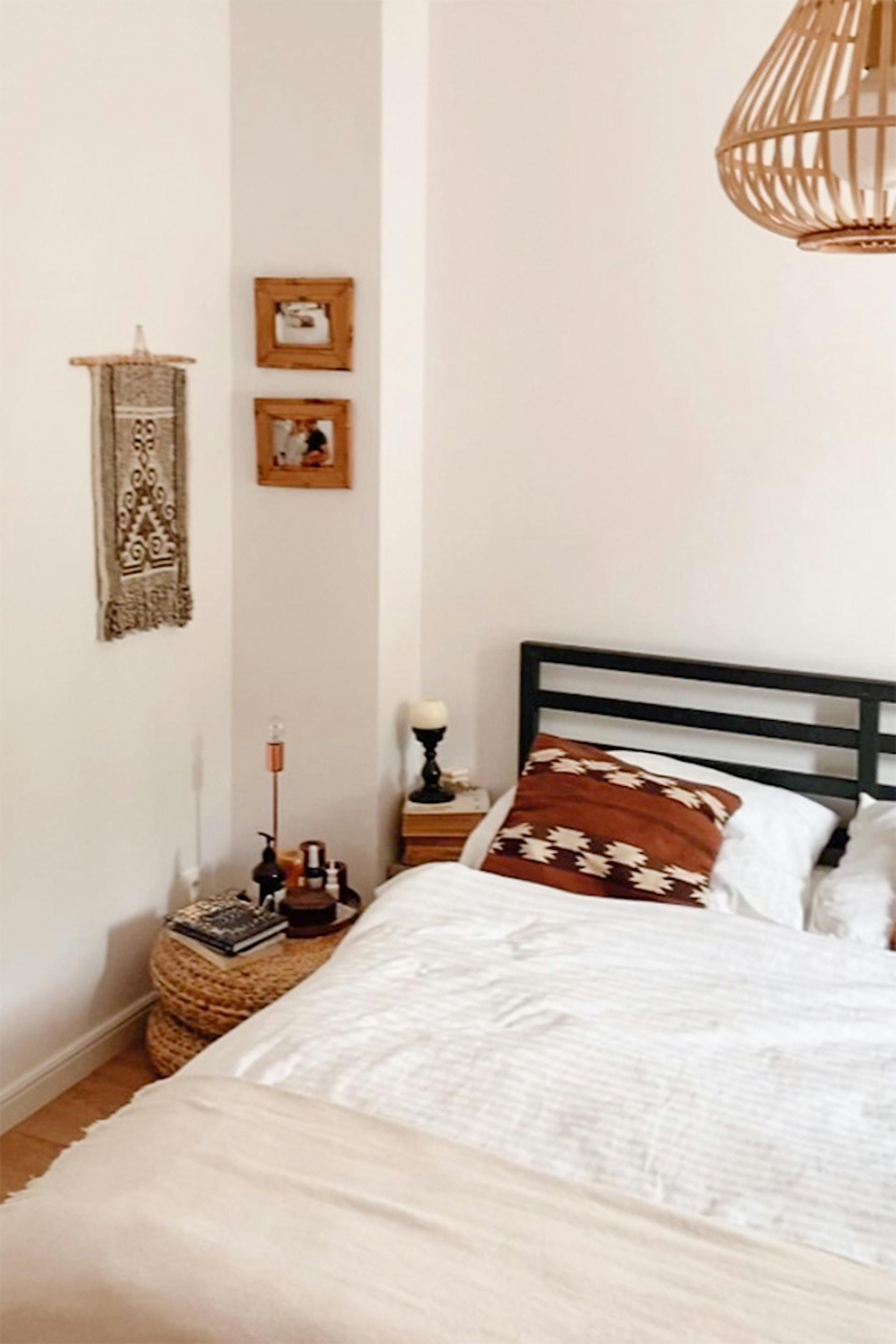 Weiße Wände in einem natürlichen Schlafzimmer, Boho-Dekorationen an einer weißen Wand im Schlafzimmer, ein Bett mit schwarzem Rahmen, gewebte Hocker, Bilder in Holzrahmen an einer weißen Wand