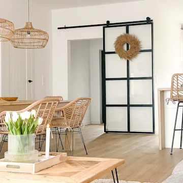 des chaises en rotin sur pieds en métal, une grande table en bois, des lampes suspendues avec un abat-jour en osier et des portes coulissantes en verre avec un cadre en métal
