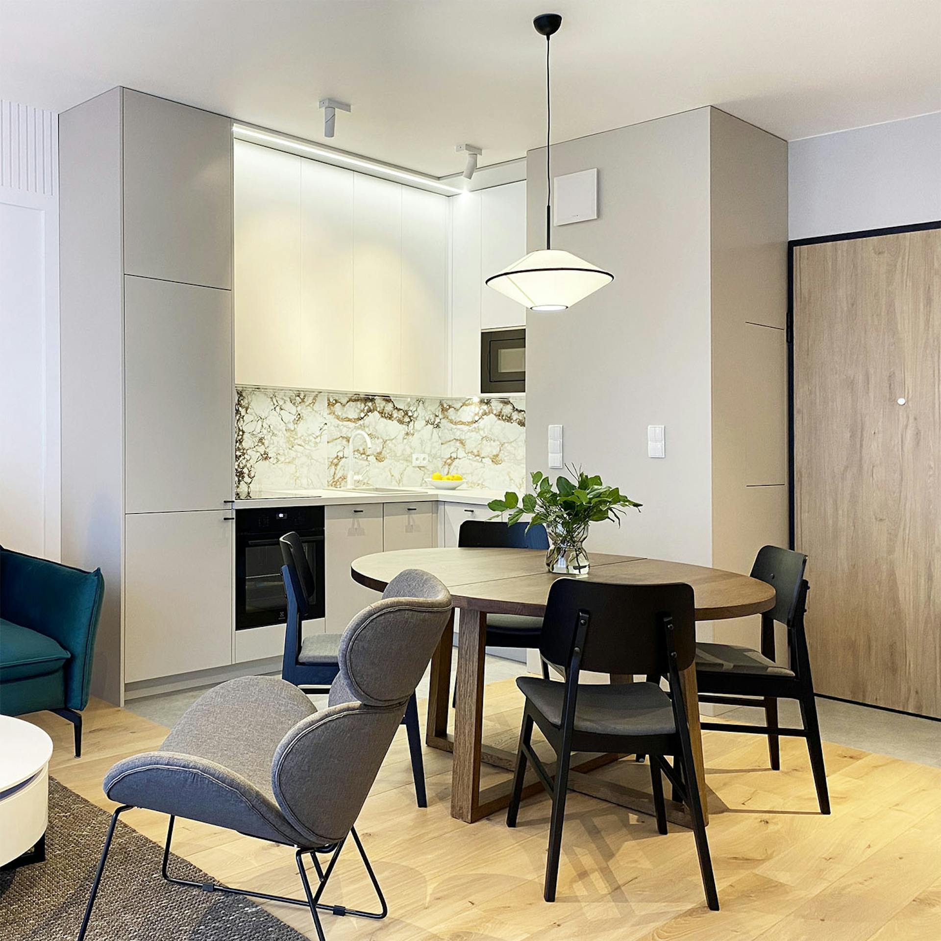 runder Esstisch in Nussbaumfarbe mit schwarzen Stühlen, hellgraue Küchenmöbel, dekorative Fliesen mit Steinmuster über der Küchenarbeitsplatte, eine Küchenzeile kombiniert mit einem Essbereich in einer modernen Wohnung