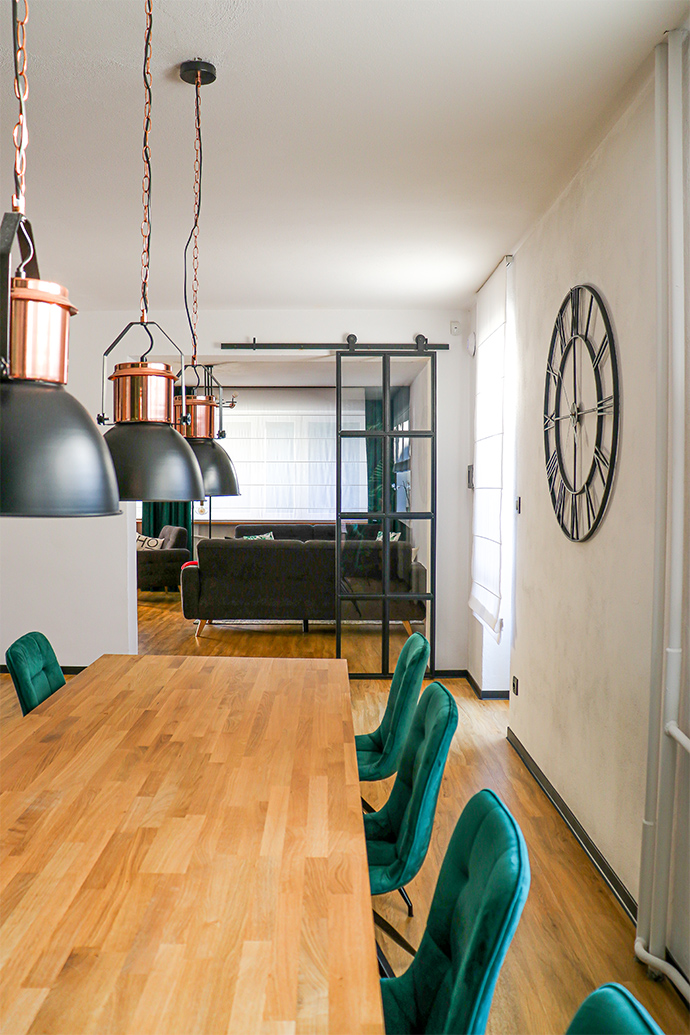 schwarze Metallwanduhr, Uhr im Industriestil, Loftdekor, Metalltür mit Glas, Lofttür, Metallglasschiebetür, schwarze Glastür, grüne Esszimmerstühle, offenes Wohnzimmer zum Esszimmer, industrielle Hängelampen für das Esszimmer, großes hölzernes Esstisch