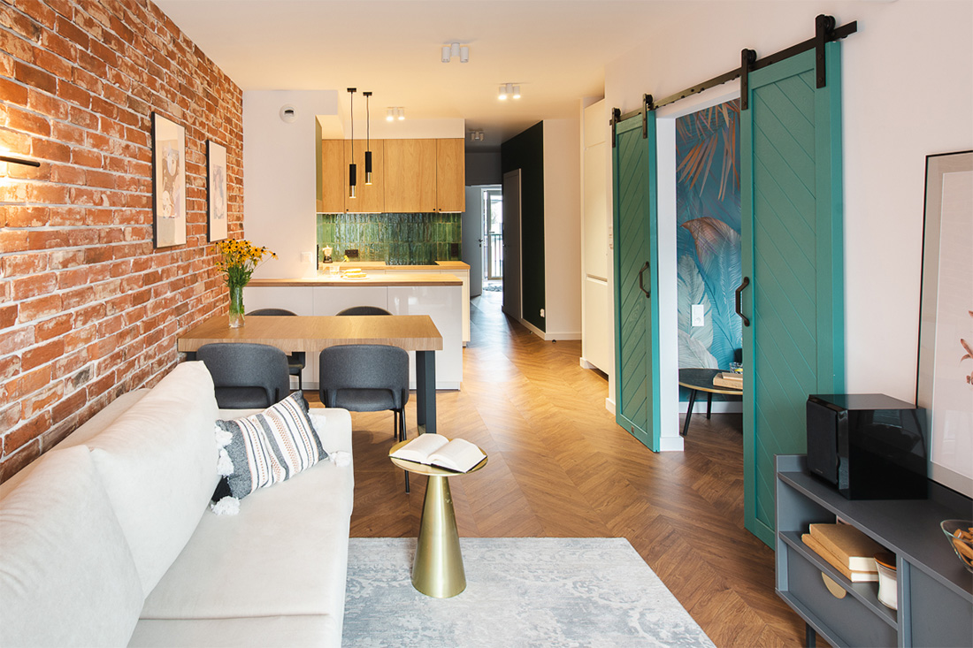 jasna sofa, czerwona cegła i turkusowe drzwi przesuwne w salonie z aneksem kuchennym