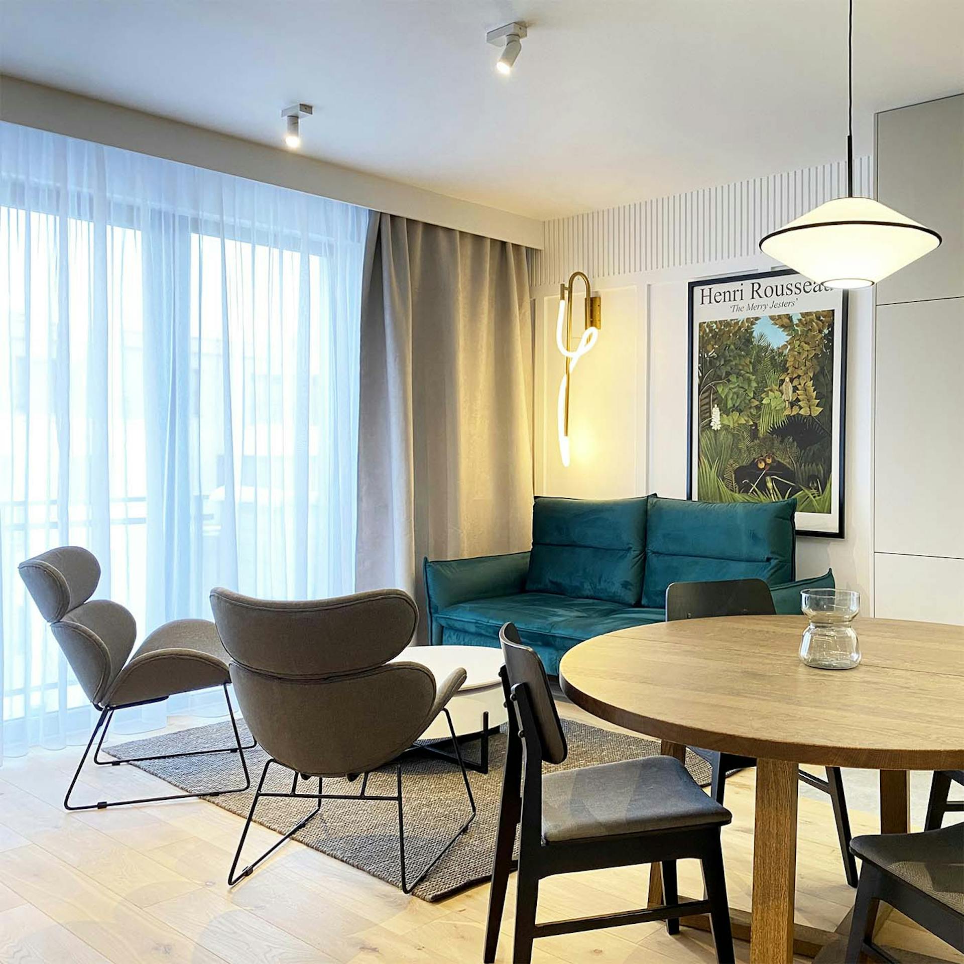 Smaragdgrünes Sofa, graue Sessel auf einem Metallgestell, Wohnzimmer kombiniert mit Essbereich in einer modernen Wohnung, runder Esstisch, moderne Beleuchtung, weiße Wände und graue Accessoires