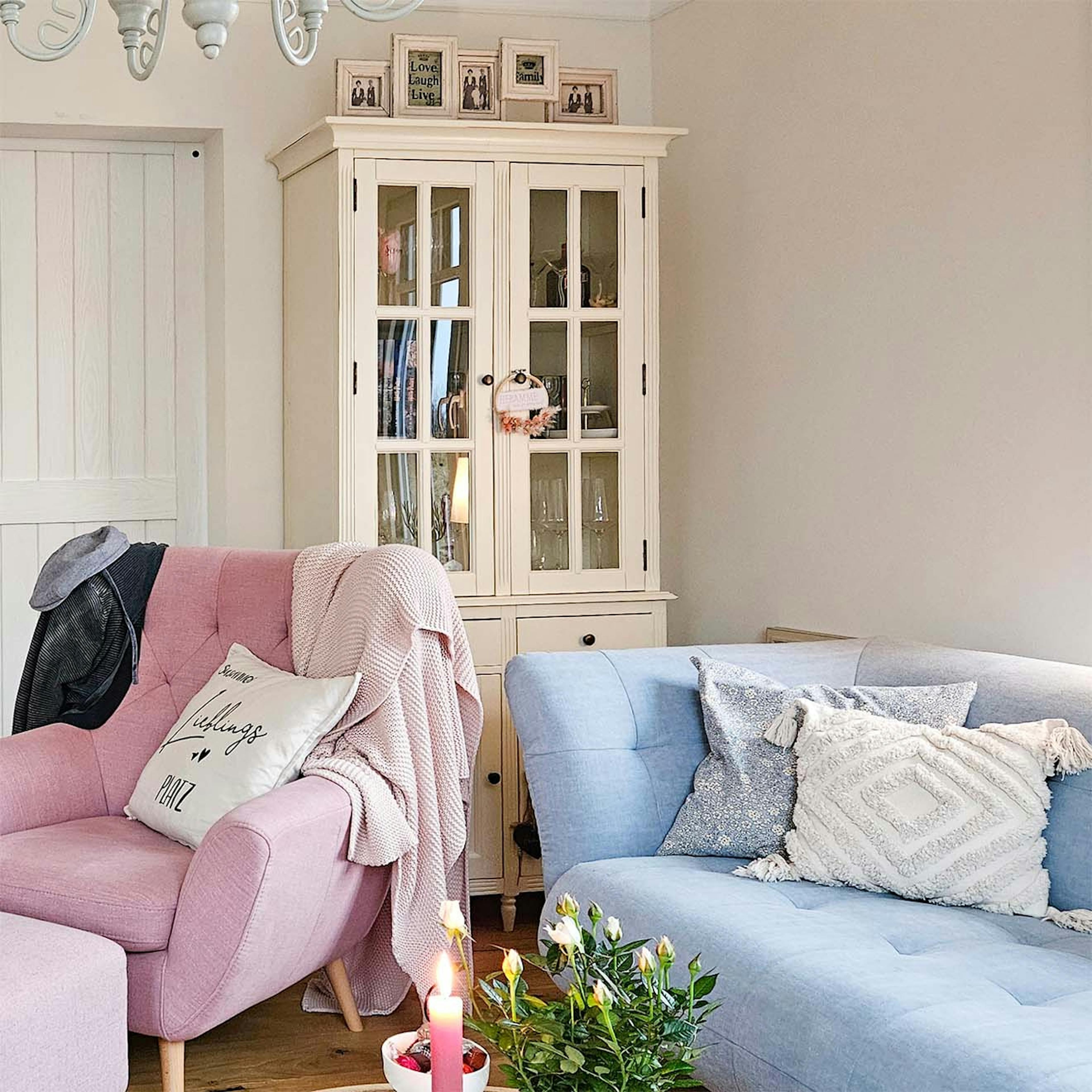 eine weiße Glasvitrine in der Ecke des Wohnzimmers, daneben ein blaues Sofa und ein rosa Sessel