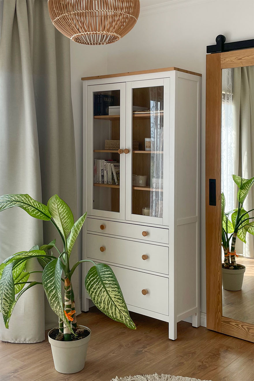 biała witryna z szufladami i szklanymi drzwiami oraz duża roślina doniczkowa w sypialni