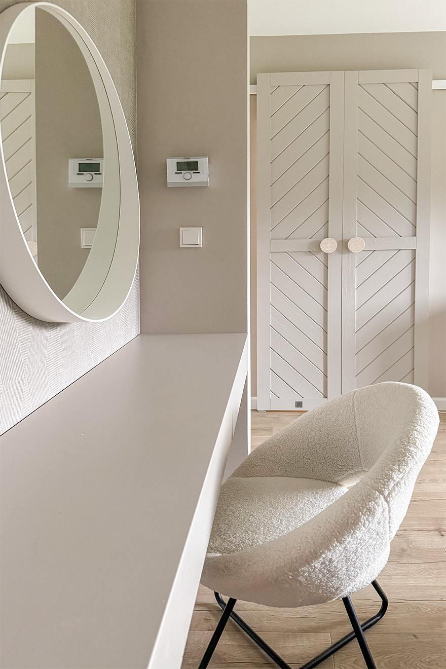 ein cremefarbener Polsterstuhl neben dem Schminktisch mit einem runden Spiegel in einem weißen Rahmen