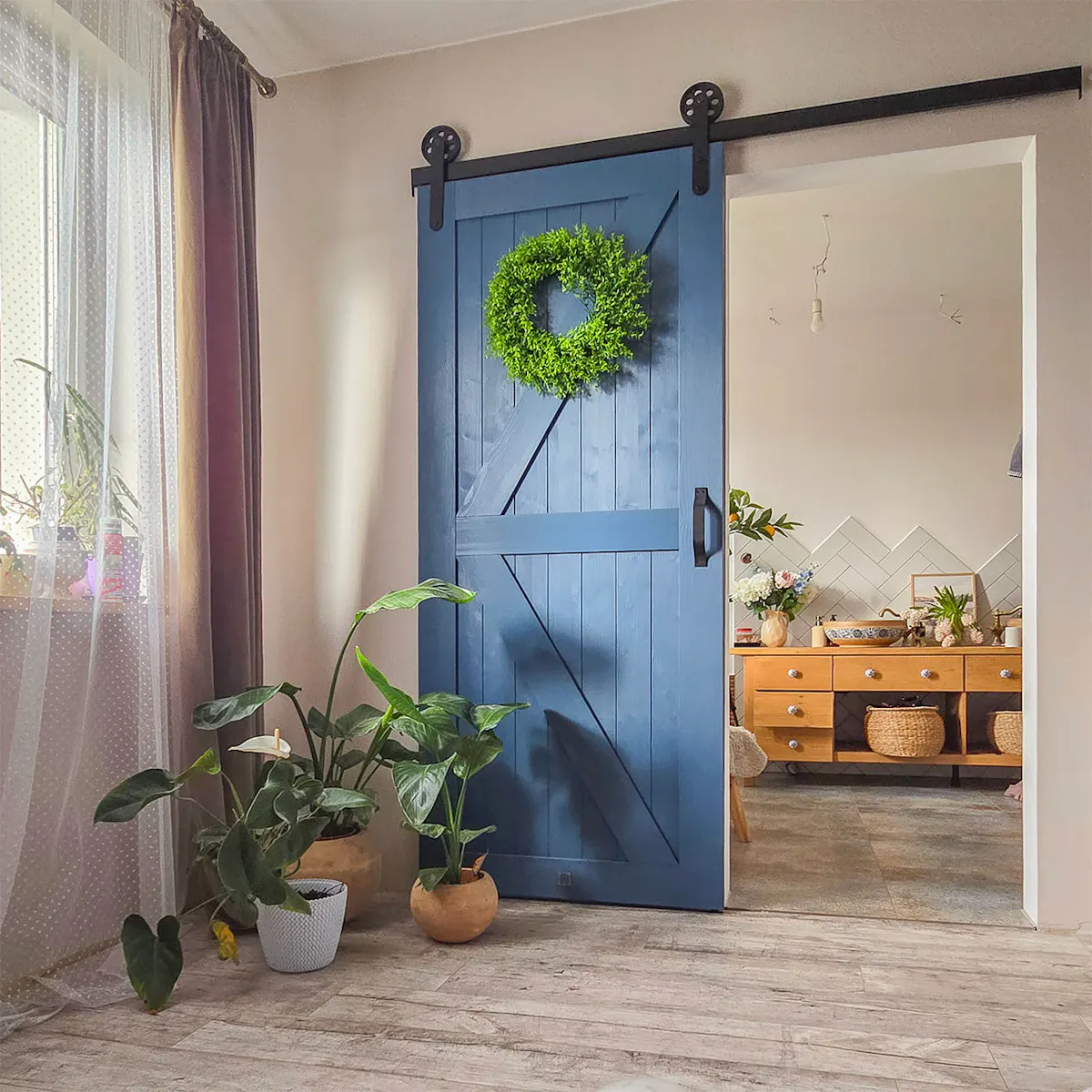 niebieskie drzwi przesuwne w stylu "barn door" łączące sypialnię z łazienką; zielony wianek na drzwiach; rośliny doniczkowe przy drzwiach