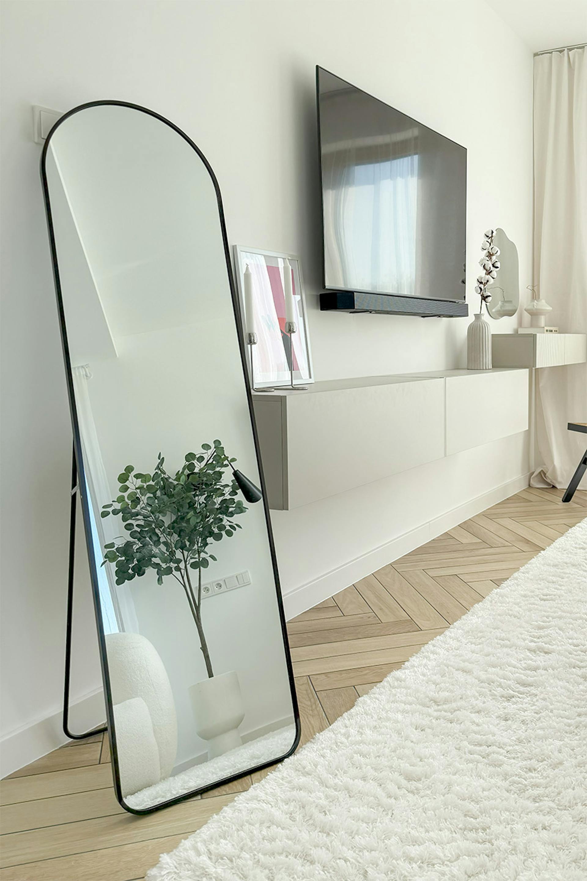 eine Ecke in einem modernen Schlafzimmer mit einem großen schwarzen gewölbten Spiegel an einer weißen Wand, einem an der Wand montierten Fernseher und einer hängenden Konsole