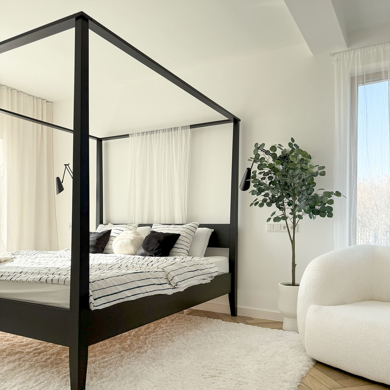 jasna sypialnia z dużym czarnym łóżkiem z baldachimem, pasiastą pościelą, dużym białym dywanem i wysoką rośliną w białej doniczce