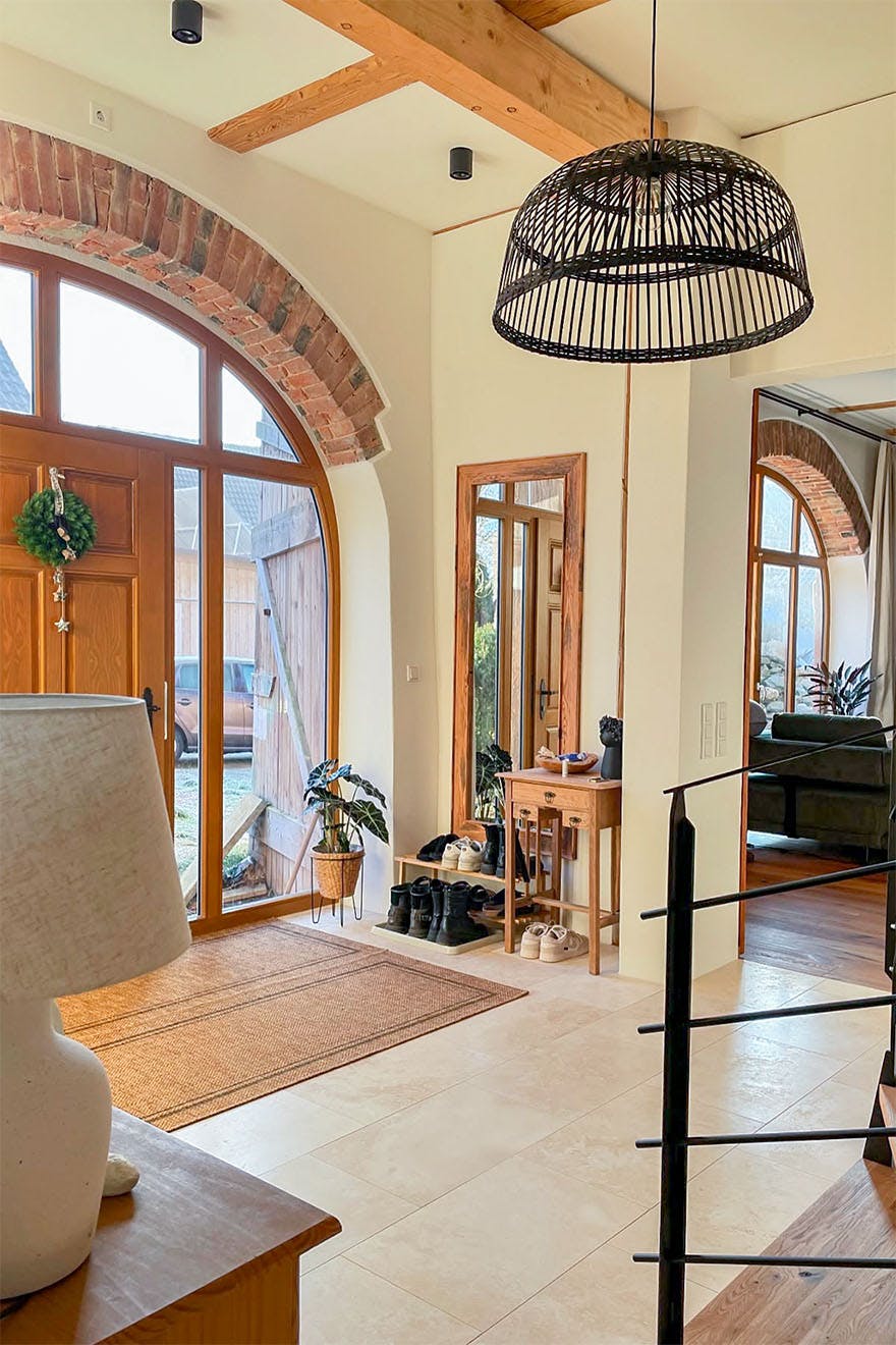 Nowoczesny hol wejściowy w stylu rustykalnym z beżowymi płytkami, dużymi łukowymi drzwiami i oknami, wiszącą lampą i drewnianymi akcentami.