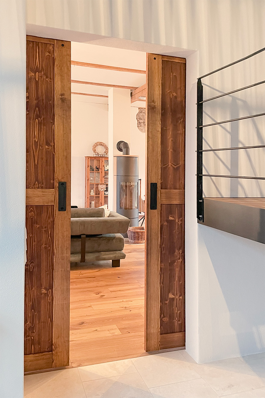 Otwarte drewniane drzwi przesuwne typu barn door, prowadzące do jasnego salonu z drewnianą podłogą, piecem wolnostojącym i ciemną sofą.