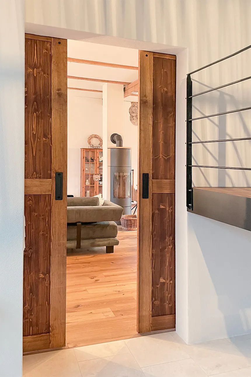 Otwarte drewniane drzwi przesuwne typu barn door, prowadzące do jasnego salonu z drewnianą podłogą, piecem wolnostojącym i ciemną sofą.