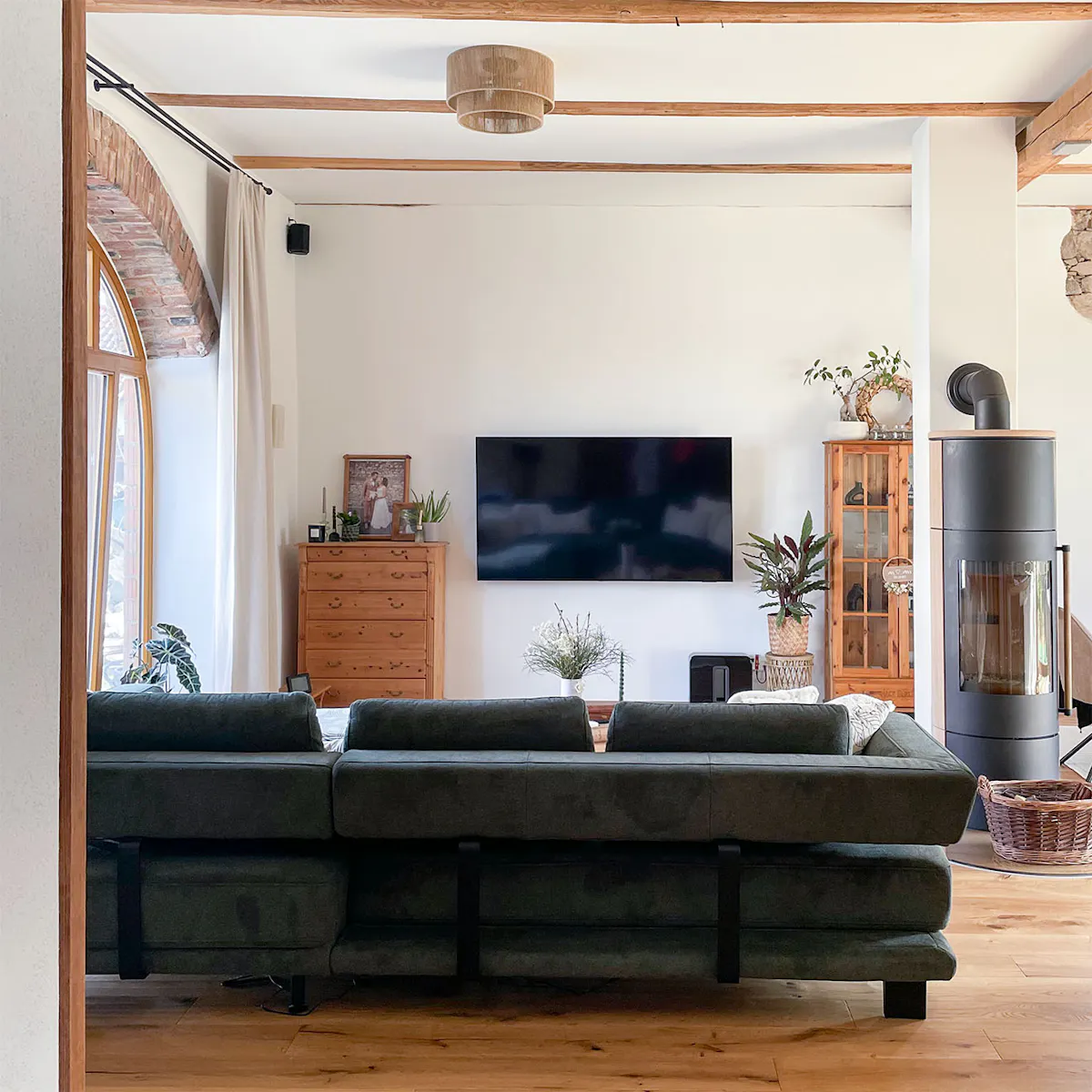 Przestronny, jasny salon w odrestaurowanej stodole, z telewizorem na ścianie, dużą ciemną sofą, drewnianymi belkami stropowymi i piecem wolnostojącym.