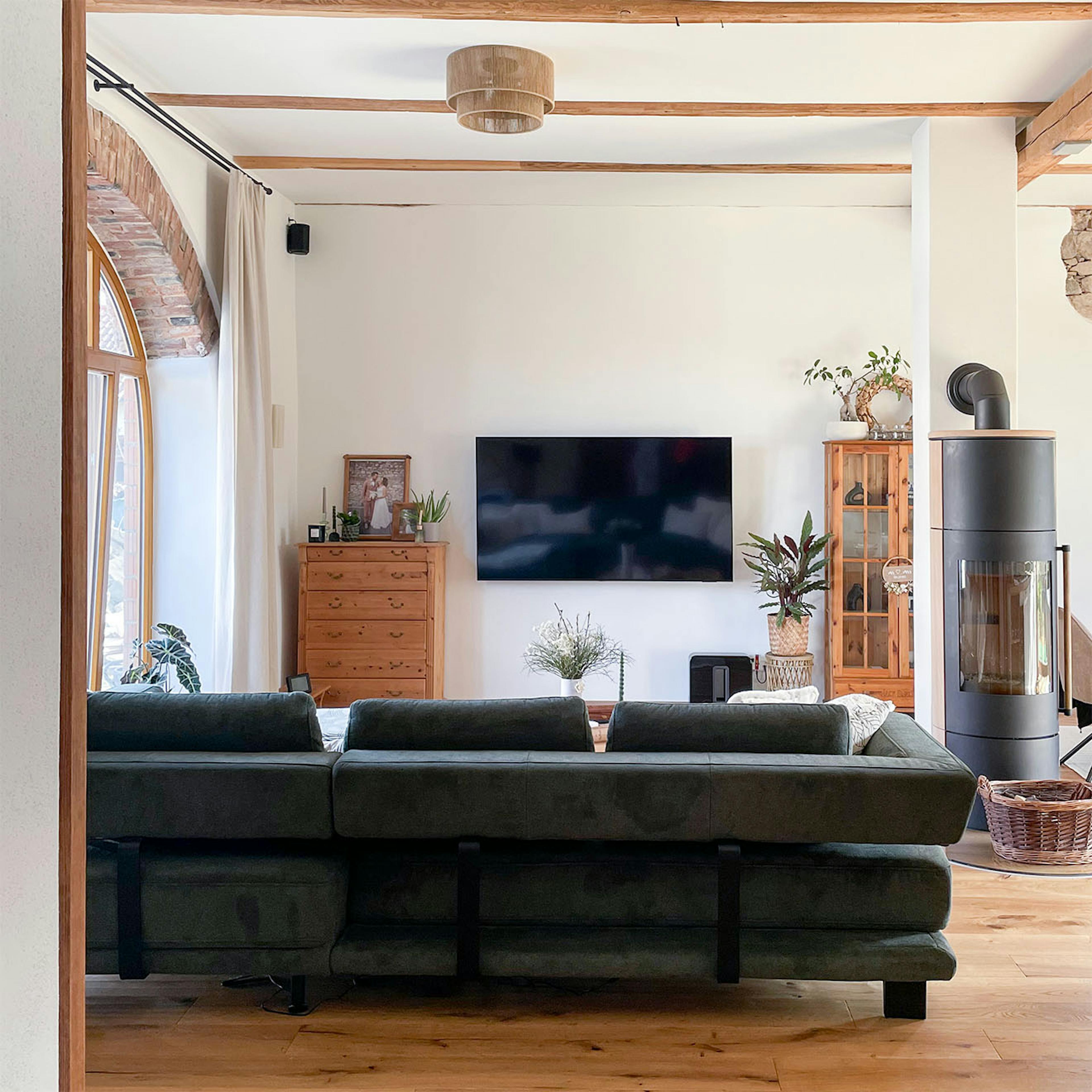 Przestronny, jasny salon w odrestaurowanej stodole, z telewizorem na ścianie, dużą ciemną sofą, drewnianymi belkami stropowymi i piecem wolnostojącym.