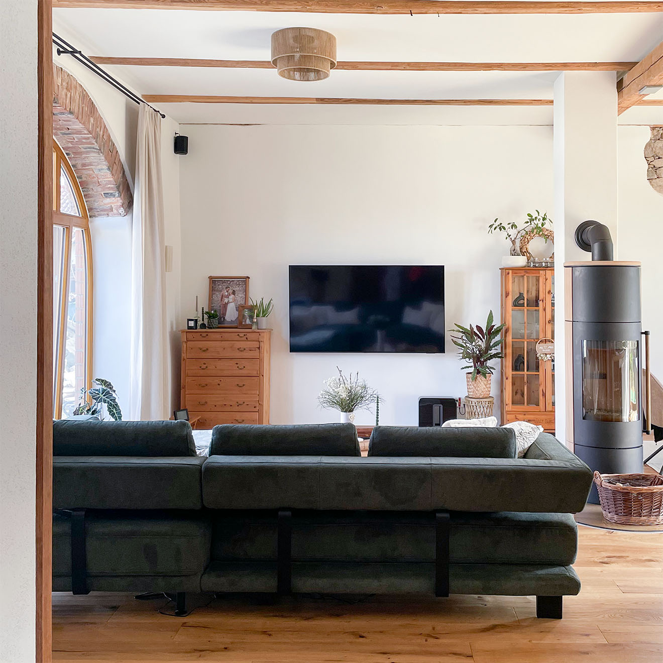 Geräumiges, helles Wohnzimmer in einem renovierten Stadl mit an der Wand montiertem Fernseher, großem dunklen Sofa, Holzbalkendecke und freistehendem Ofen.