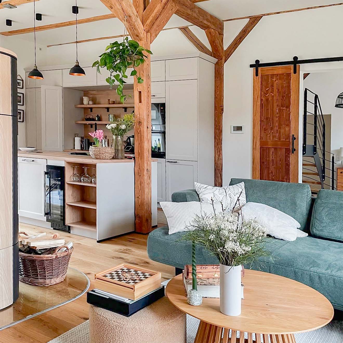 Offener Wohn- und Küchenbereich in einem renovierten Stadl, mit sichtbaren Holzbalken, einem grünen Sofa, modernen weißen Küchenmöbeln und rustikalen Details.
