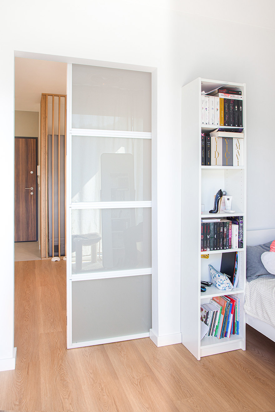 Białe przesuwne drzwi ze szkłem mlecznym, oddzielające sypialnię z regałem na książki od korytarza z drewnianymi lamelami.