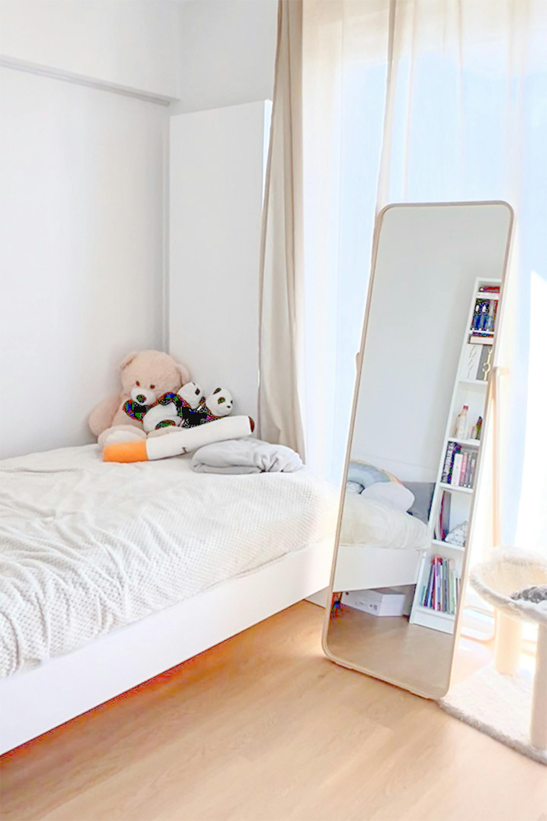 Helles Schlafzimmer mit einem weißen Bett, auf dem Plüschbären liegen, sowie einem großen, bodenstehenden Spiegel, der auf dem Holzboden am Fenster steht.