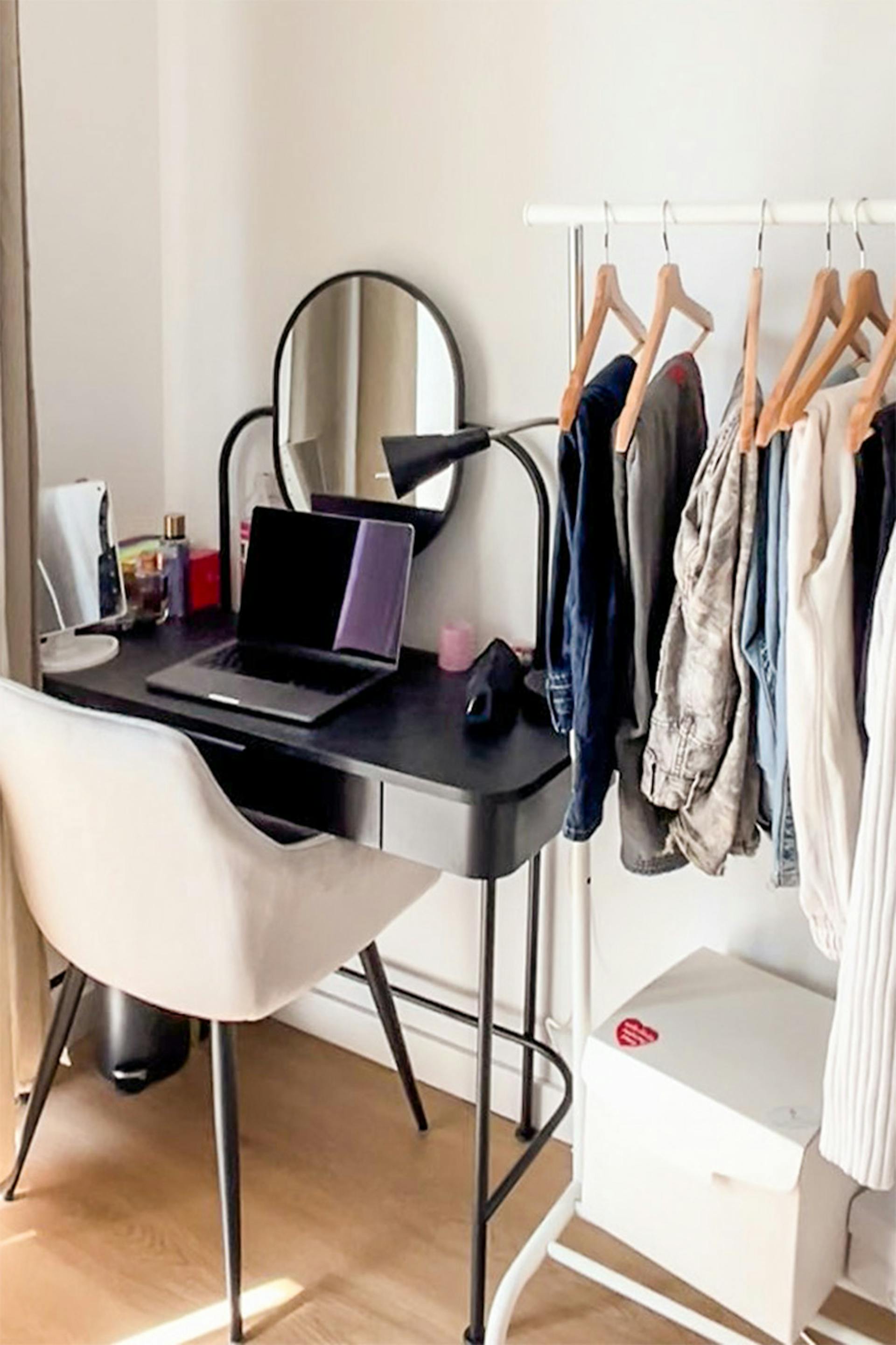 Schwarzer Schreibtisch mit rundem Spiegel, daneben ein gepolsterter Stuhl und ein stehender Kleiderständer in einem Raum mit hellen Wänden und Holzboden.