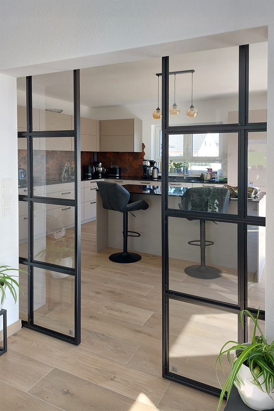 Przejście z salonu do przestronnej kuchni przez czarne industrialne drzwi. Widoczna kuchnia z wyspą, beżowymi frontami i miedzianymi płytkami.