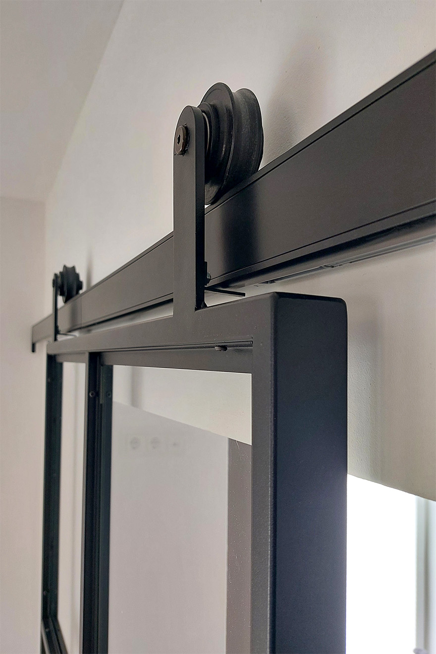 Zbliżenie na mechanizm przesuwny czarnych, metalowo-szklanych drzwi industrialnych, widoczna prowadnica i rolka.