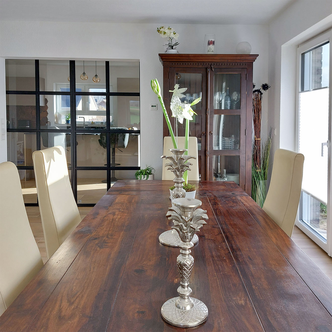 Drewniany stół w jadalni z dekoracjami, w tle klasyczna witryna i czarne industrialne drzwi prowadzące do kuchni.