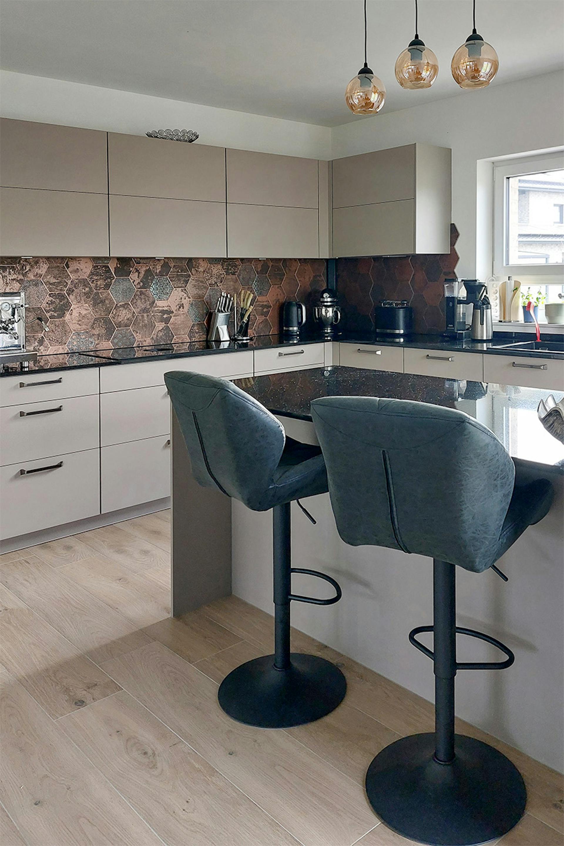 Kuchnia z wyspą, beżowe fronty, miedziane płytki, czarne hokery i wiszące lampy - nowoczesny i funkcjonalny design.