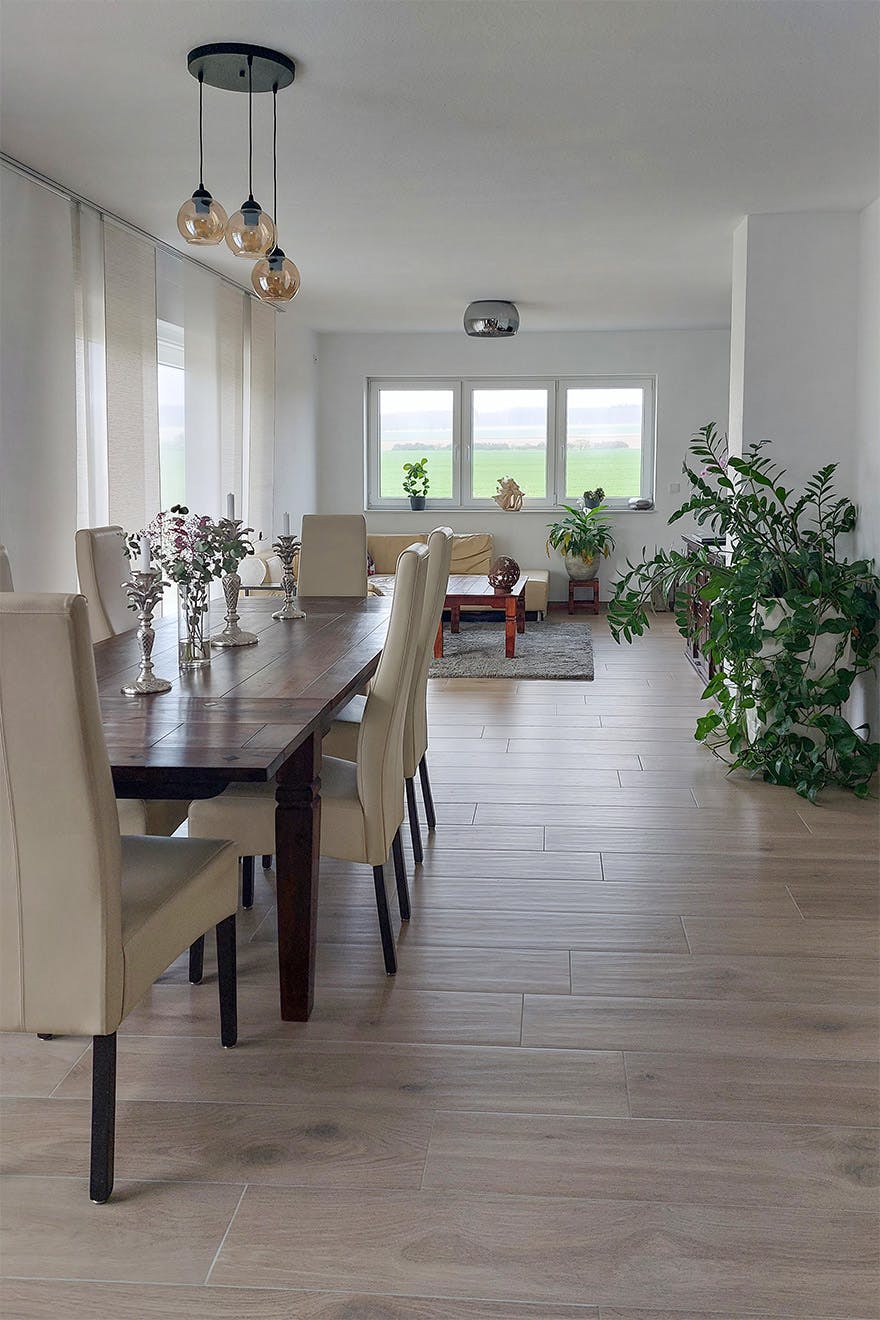 Ein helles Esszimmer mit einem langen Holztisch und cremefarbenen Stühlen, im Hintergrund ein großes Wohnzimmer mit einem beigen Sofa und Fenstern mit Blick ins Grüne.