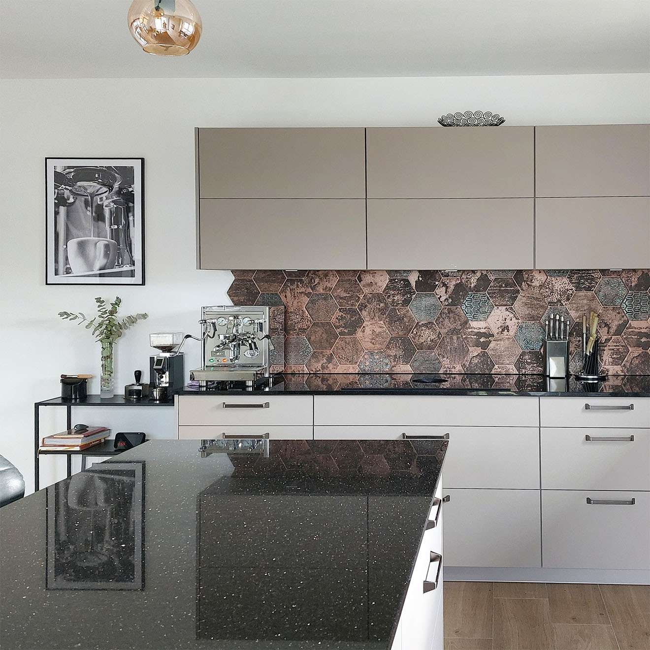 Moderne Küche mit beigen Schränken, dunkler Arbeitsplatte, sechseckigen Kupferfliesen und einer silbernen Kaffeemaschine.