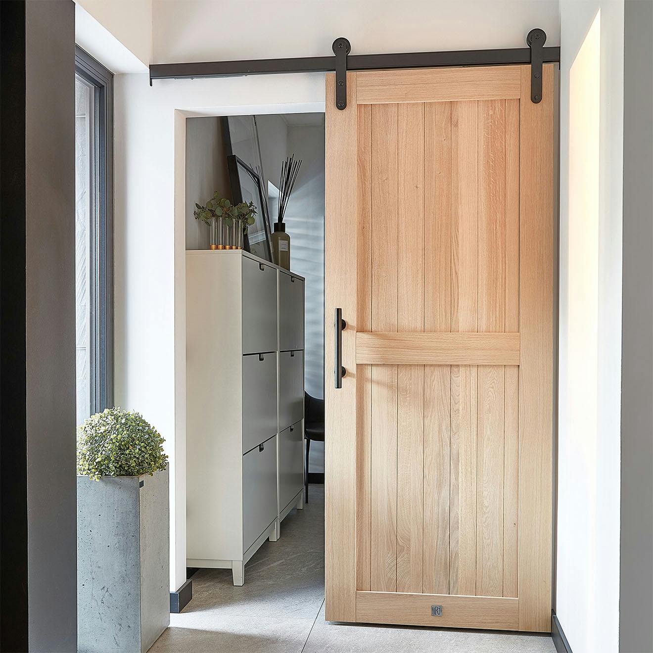 Elegancki wiatrołap z drewnianymi drzwiami przesuwnymi prowadzącymi do garderoby. Drzwi z naturalnego dębu z czarnym okuciem, duża doniczka z rośliną i szafka na buty tworzą funkcjonalną i stylową przestrzeń.