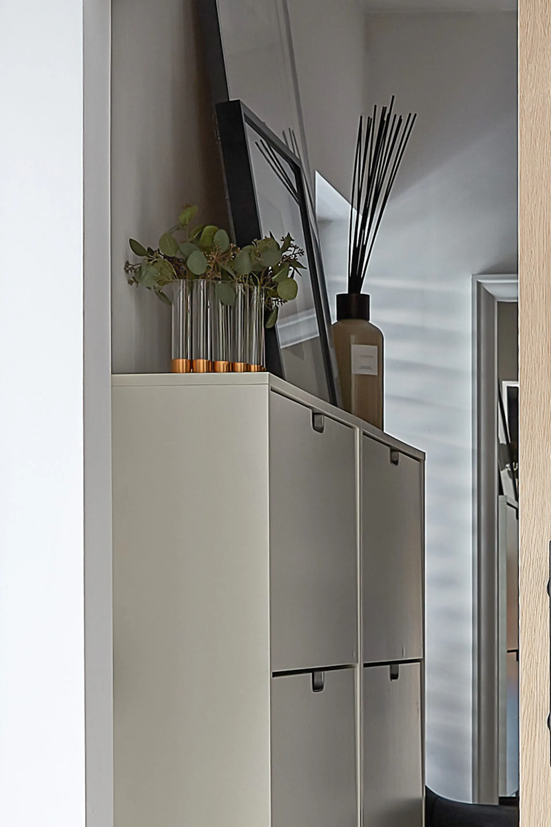 Detale wnętrza garderoby, widoczny przez uchylone dębowe drzwi. Na białej szafce na buty stoją stylowe dekoracje: wazon z trawą i dyfuzor zapachowy, co nadaje wnętrzu przytulności i elegancji.