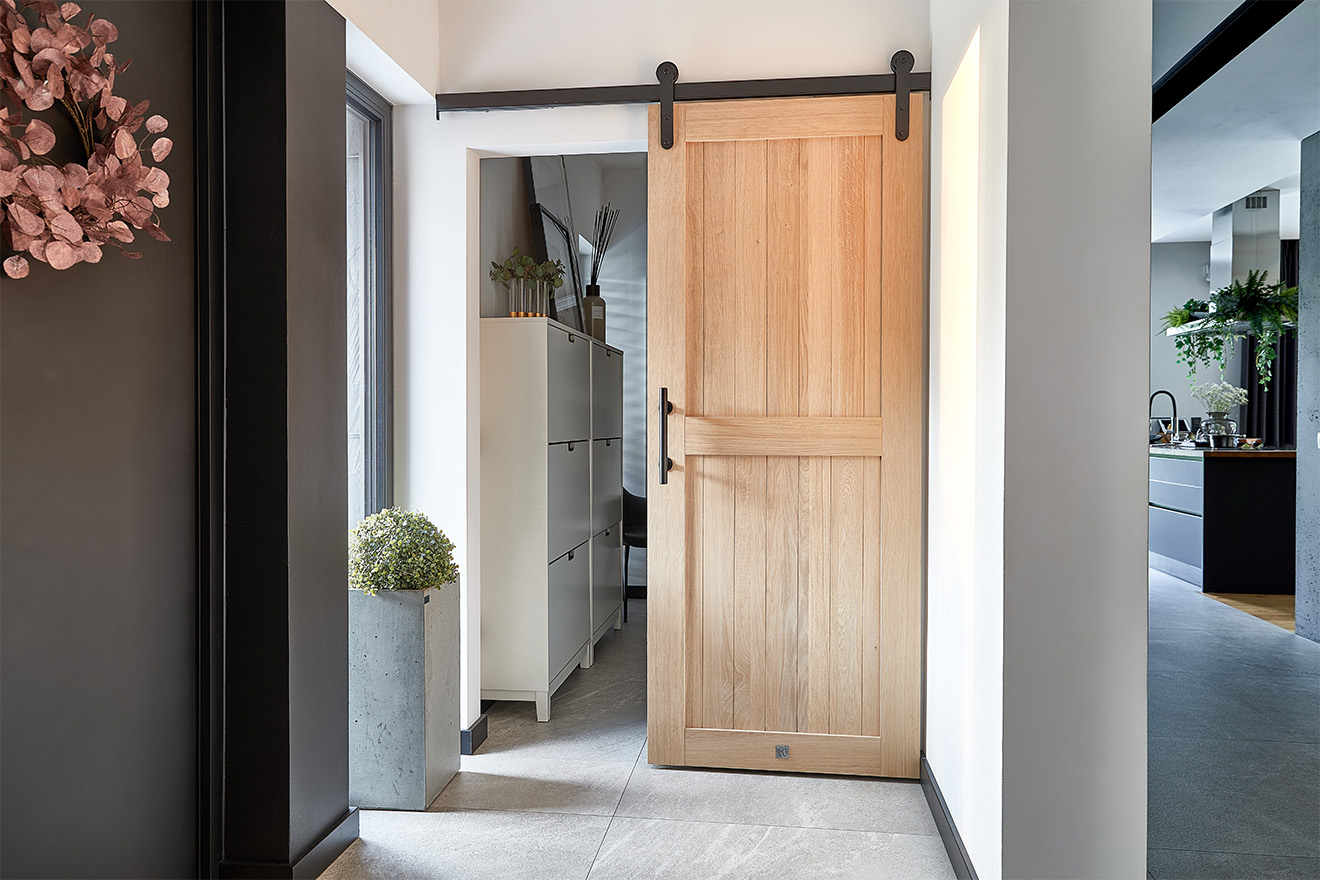 Schiebetüren aus Eichenholz in einem modernen Windfang. Eine helle, hölzerne Tür im Scheunenstil, die an einer schwarzen Schiene hängt, öffnet sich zu einem begehbaren Ankleide mit einem weißen Schuhschrank.